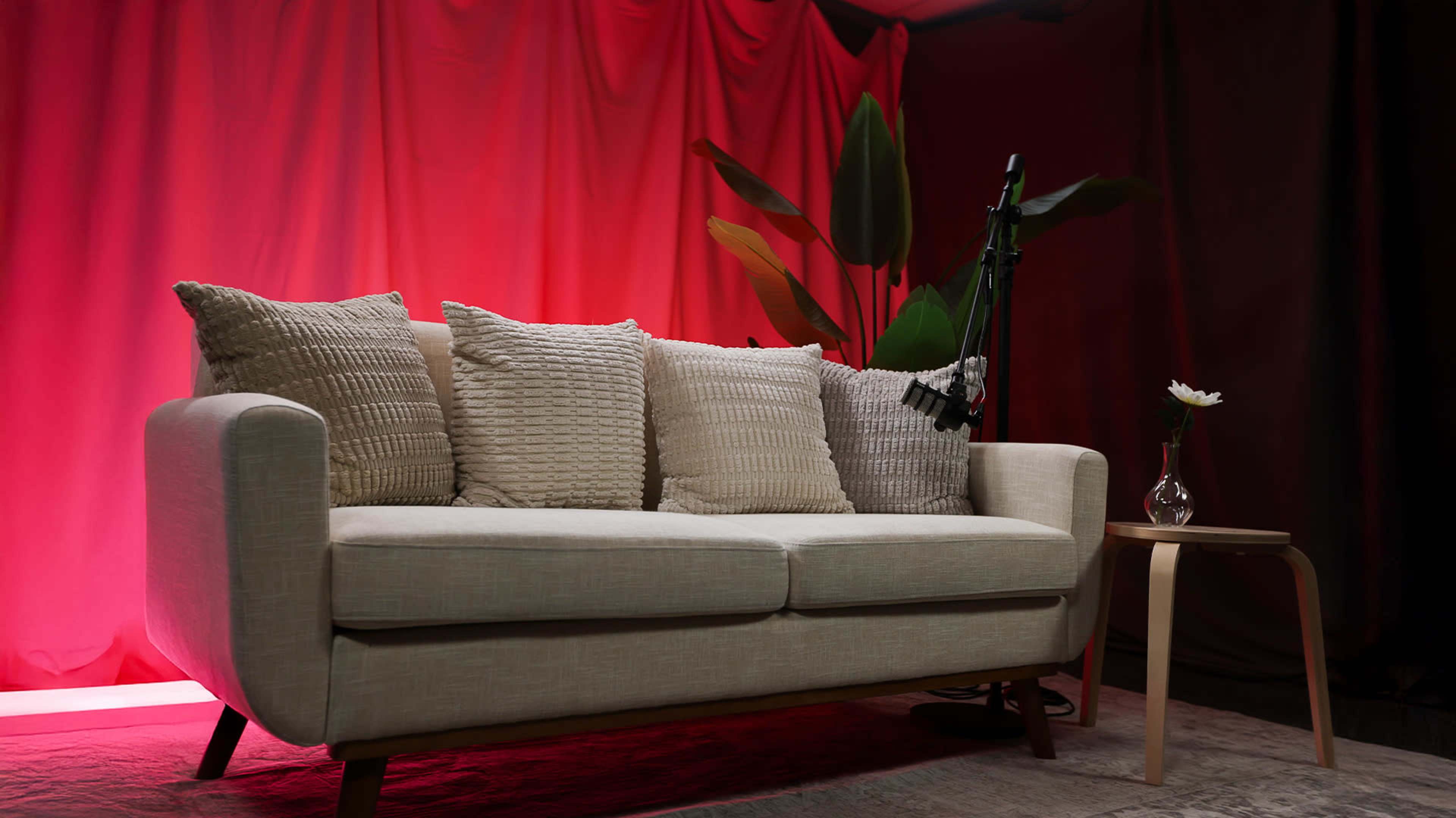 A light-colored sofa with decorative pillows is positioned next to a small table holding a vase with a flower, in front of a bright red backdrop.