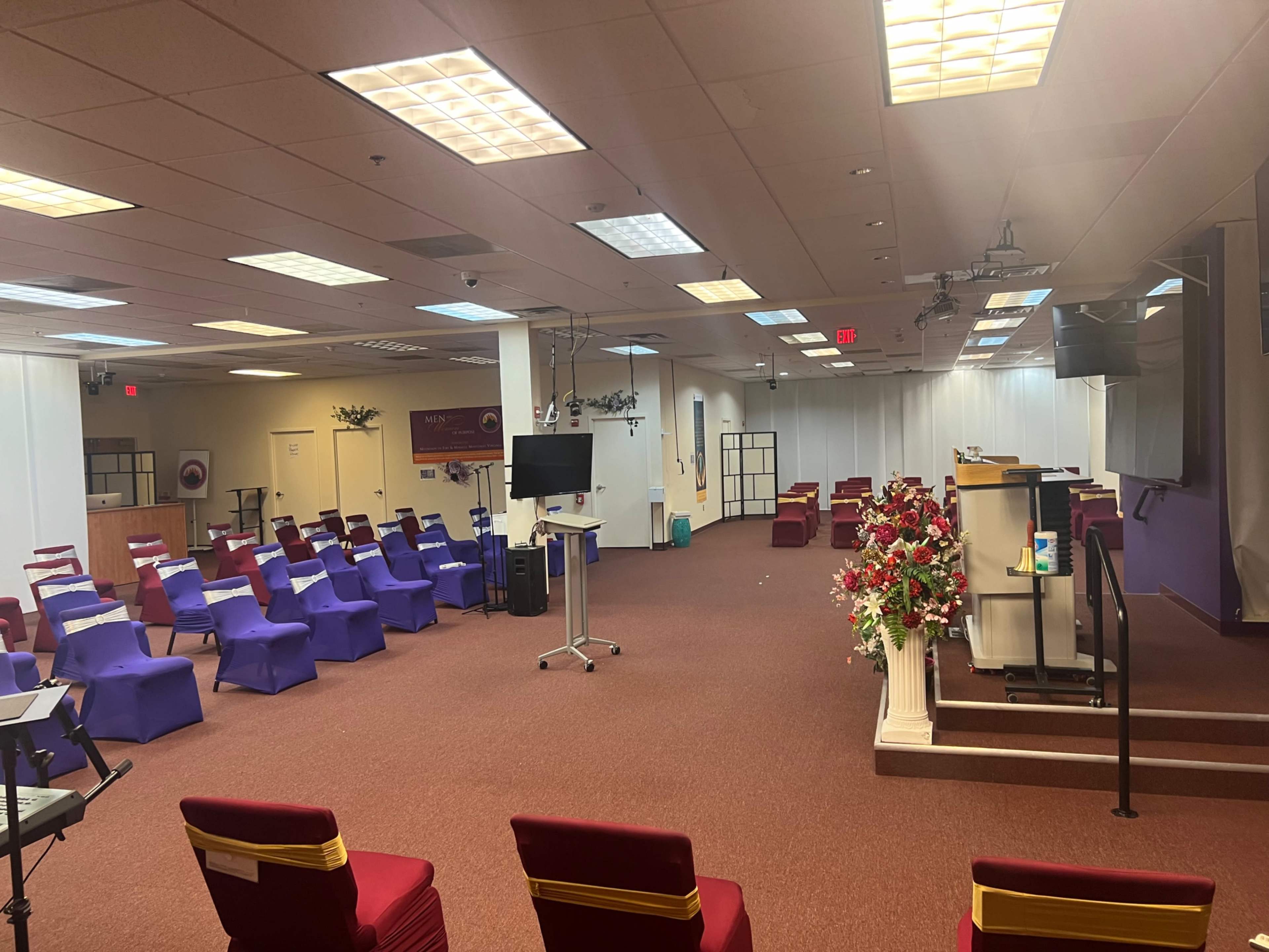 The image shows a spacious room set up with rows of purple and red chairs facing a podium and decorative flowers on a stand.