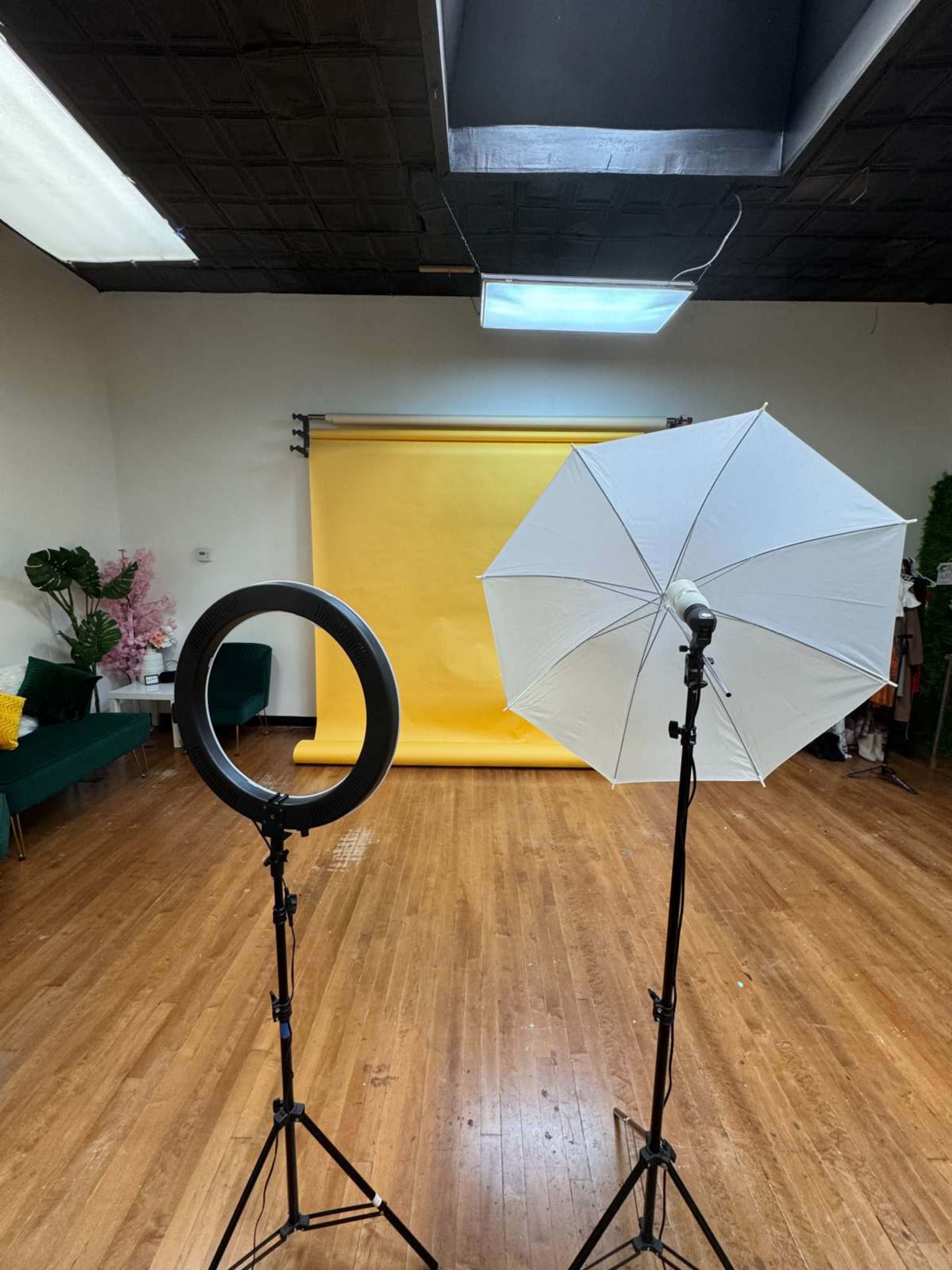 The image shows a photography studio with a yellow backdrop, two lighting setups—one with a circular softbox and another with a reflective umbrella—positioned on either side.