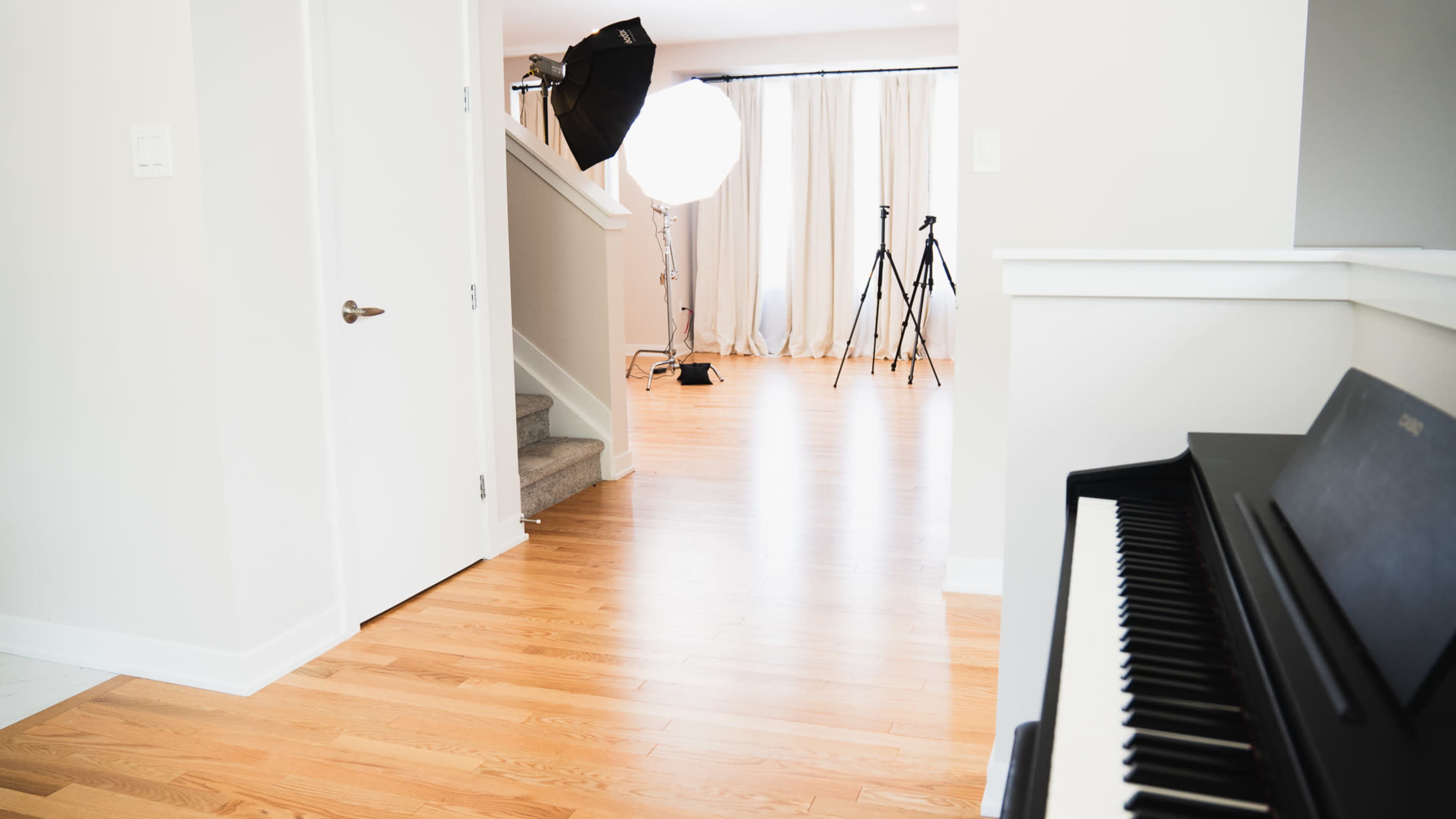 A well-lit interior space featuring a staircase, a piano in the foreground, and photography equipment, including a softbox and tripods, set up in the background.