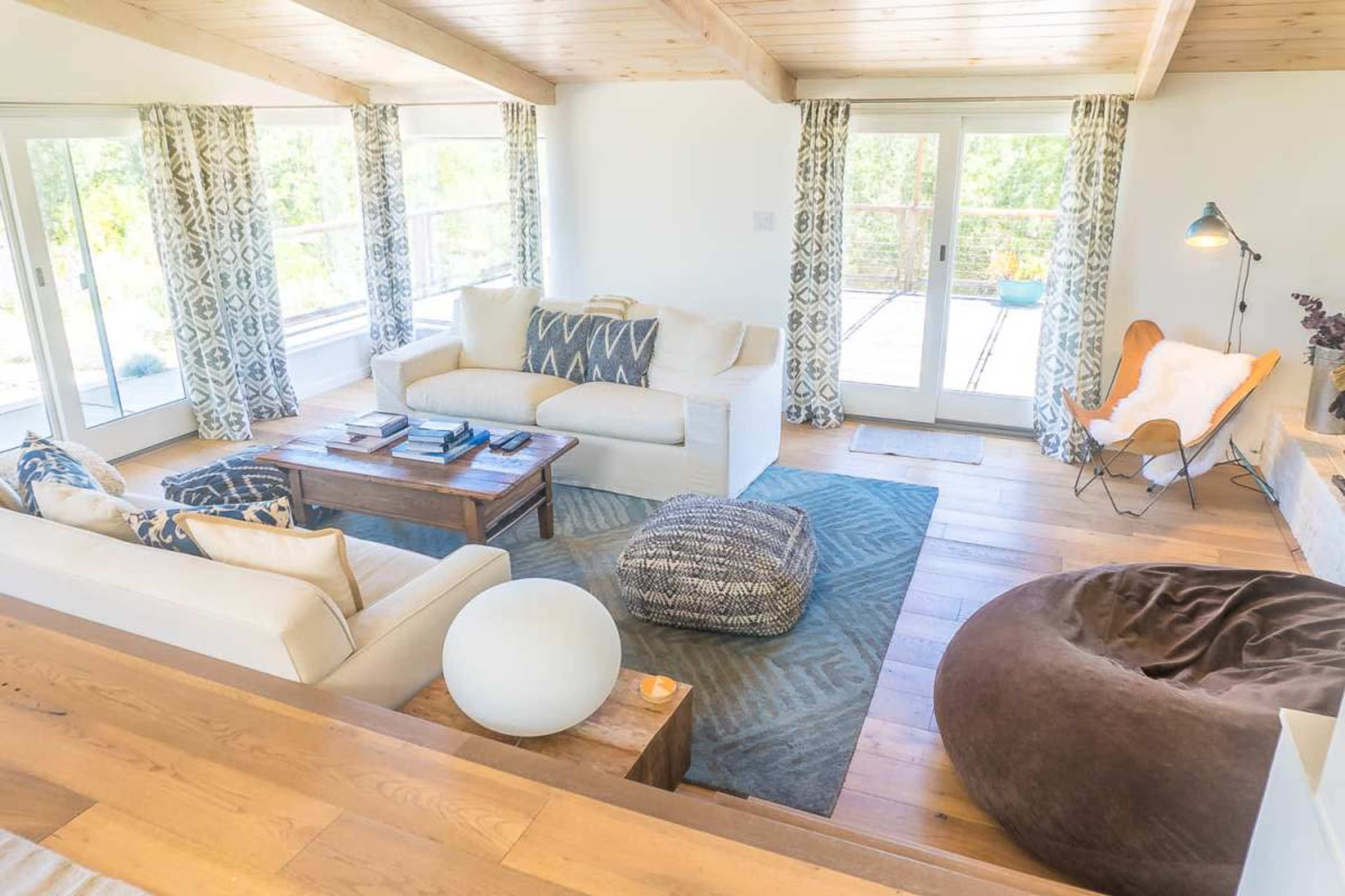 The image shows a bright living room with large windows, a light-colored sofa, a wooden coffee table, and various seating options including a bean bag and a butterfly chair.