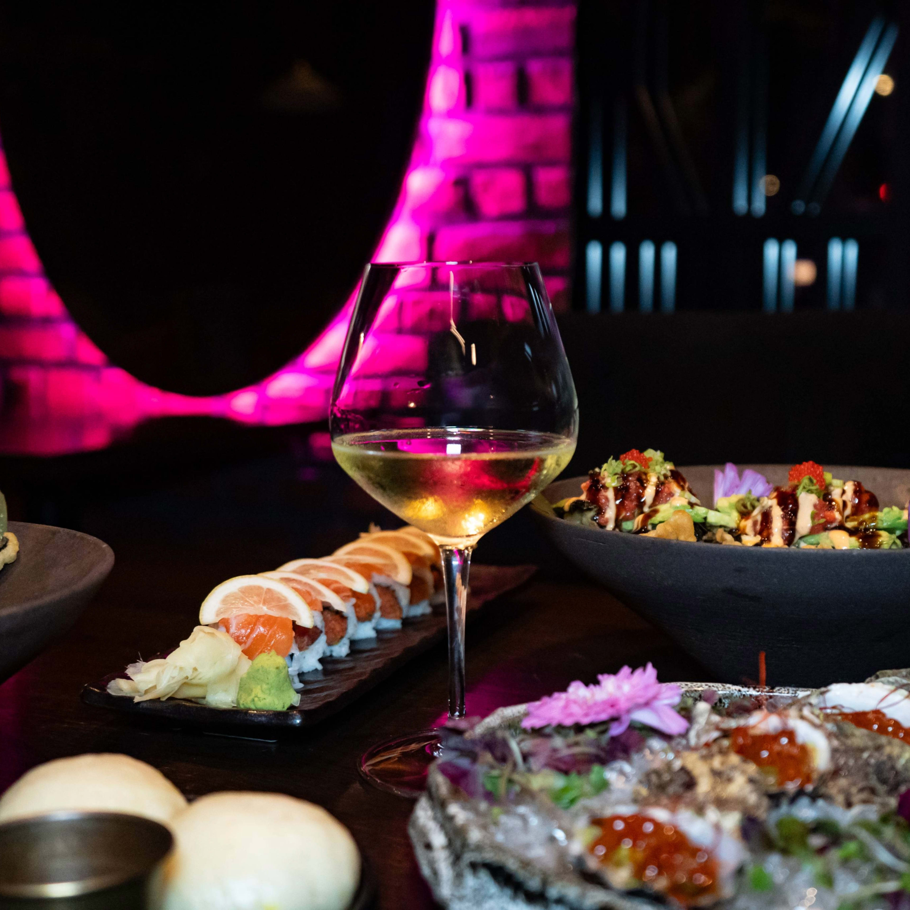 The image showcases a table setting with sushi, a glass of white wine, and colorful dishes in a dimly lit restaurant featuring a pink illuminated backdrop.