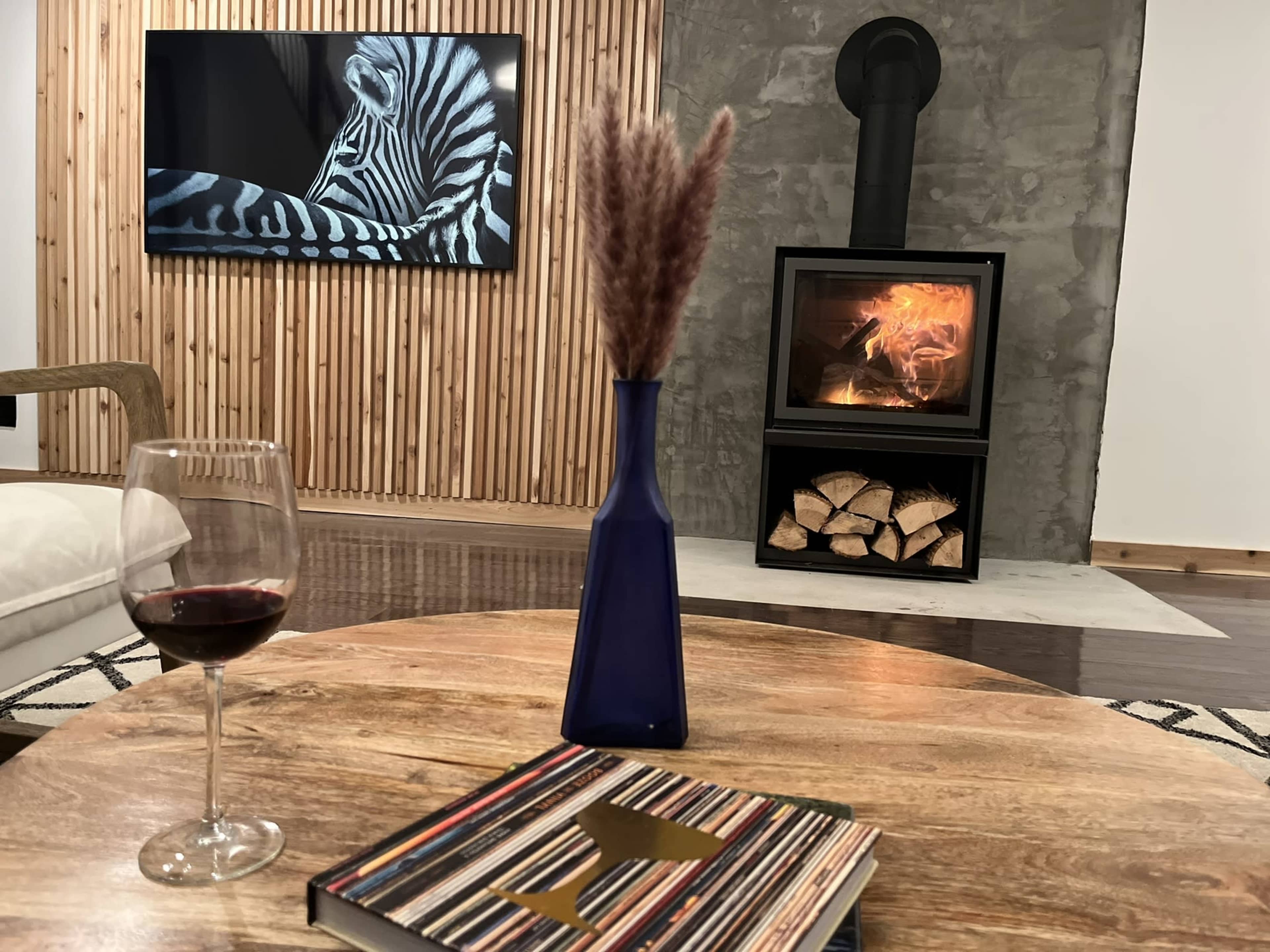A cozy living room features a glass of red wine on a wooden table, a vase with dry grass, a modern wood stove with logs stacked beside it, and a zebra photograph displayed on the wall.