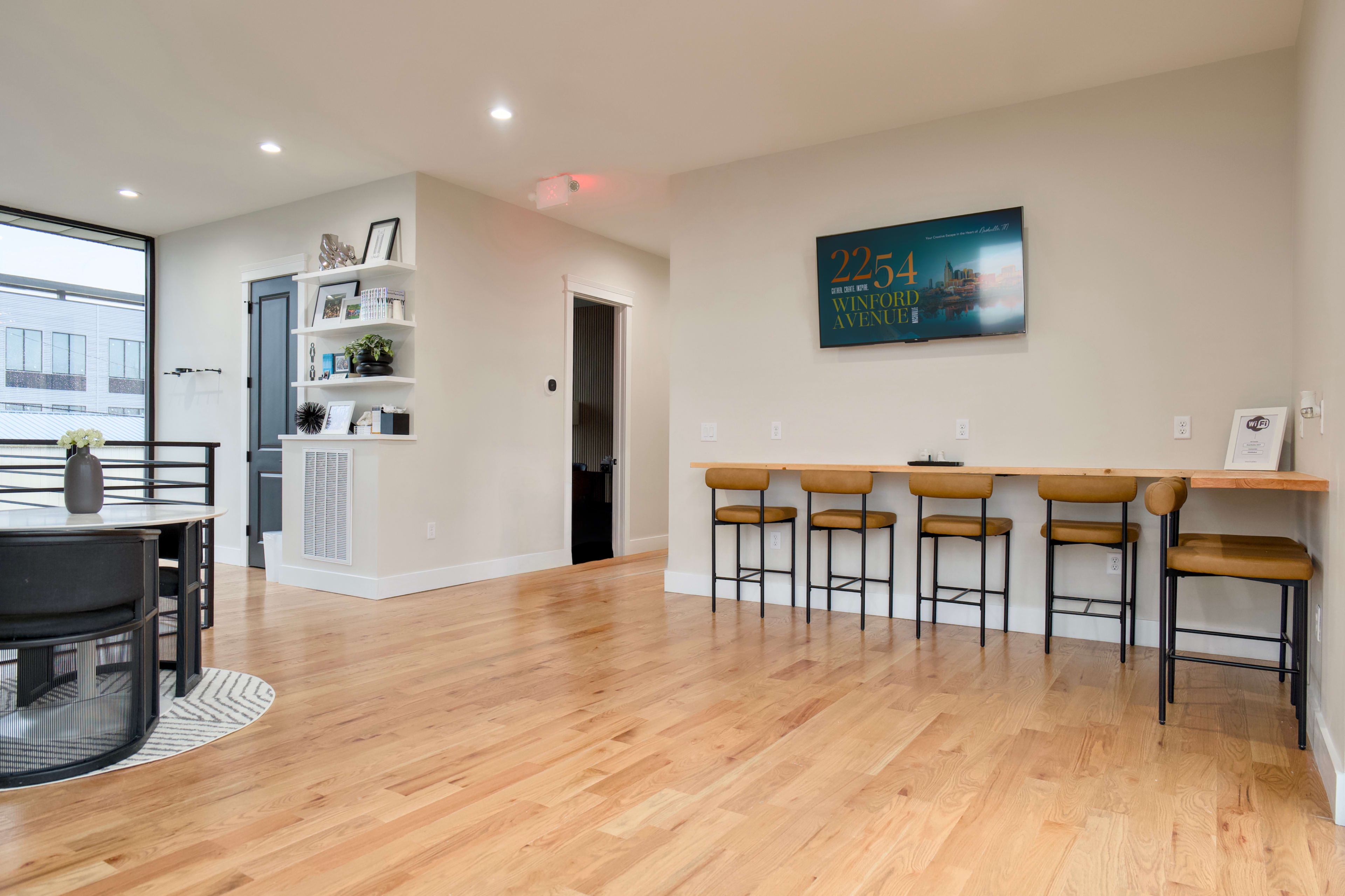 The image shows a modern interior space with wooden flooring, a dining area with a round table, a kitchen bar with stools, and a minimalist decor featuring a wall-mounted shelf and a framed sign.