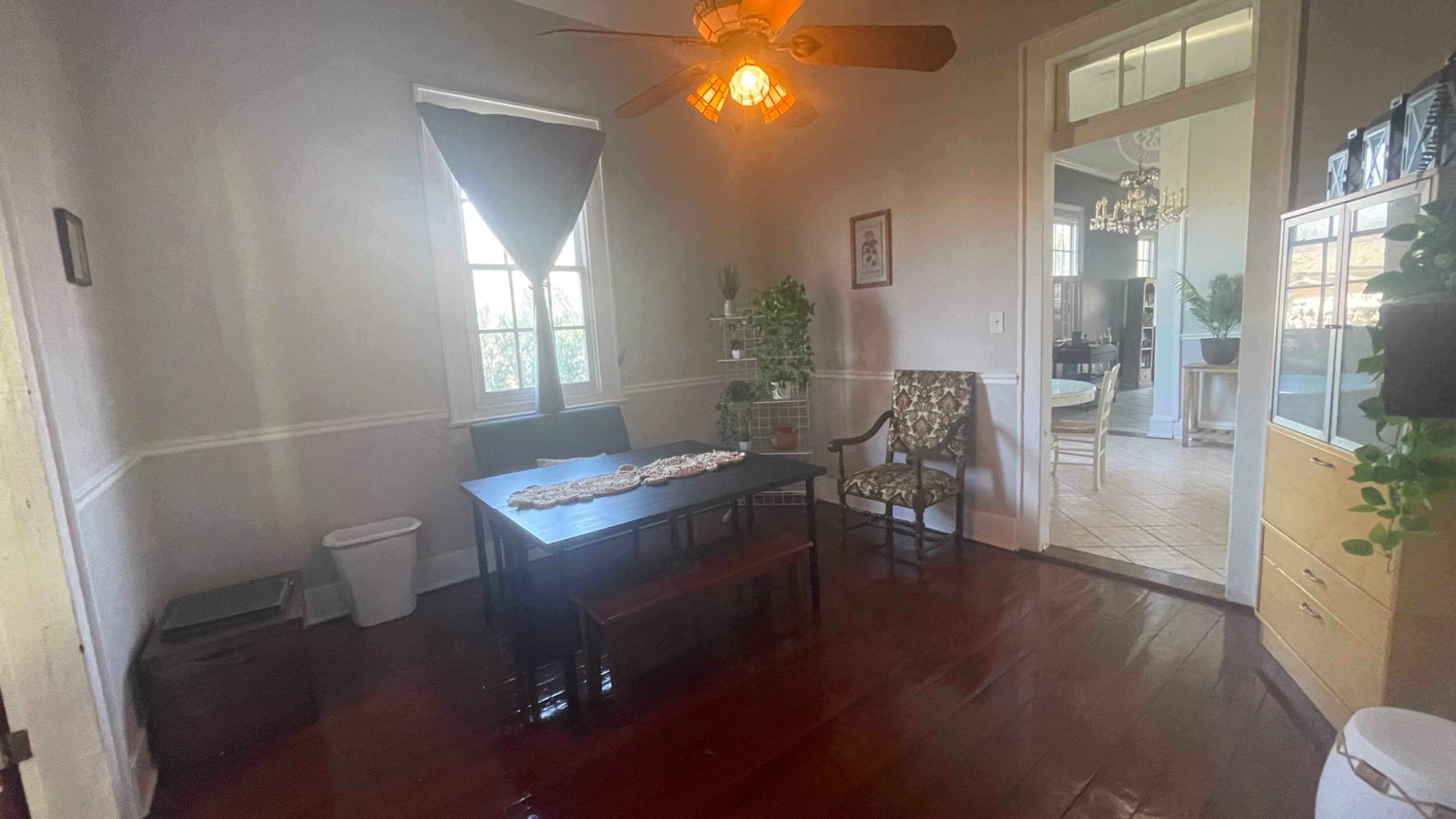 The image shows a well-lit dining room with a wooden table and chairs, a ceiling fan, and a view into an adjoining room.