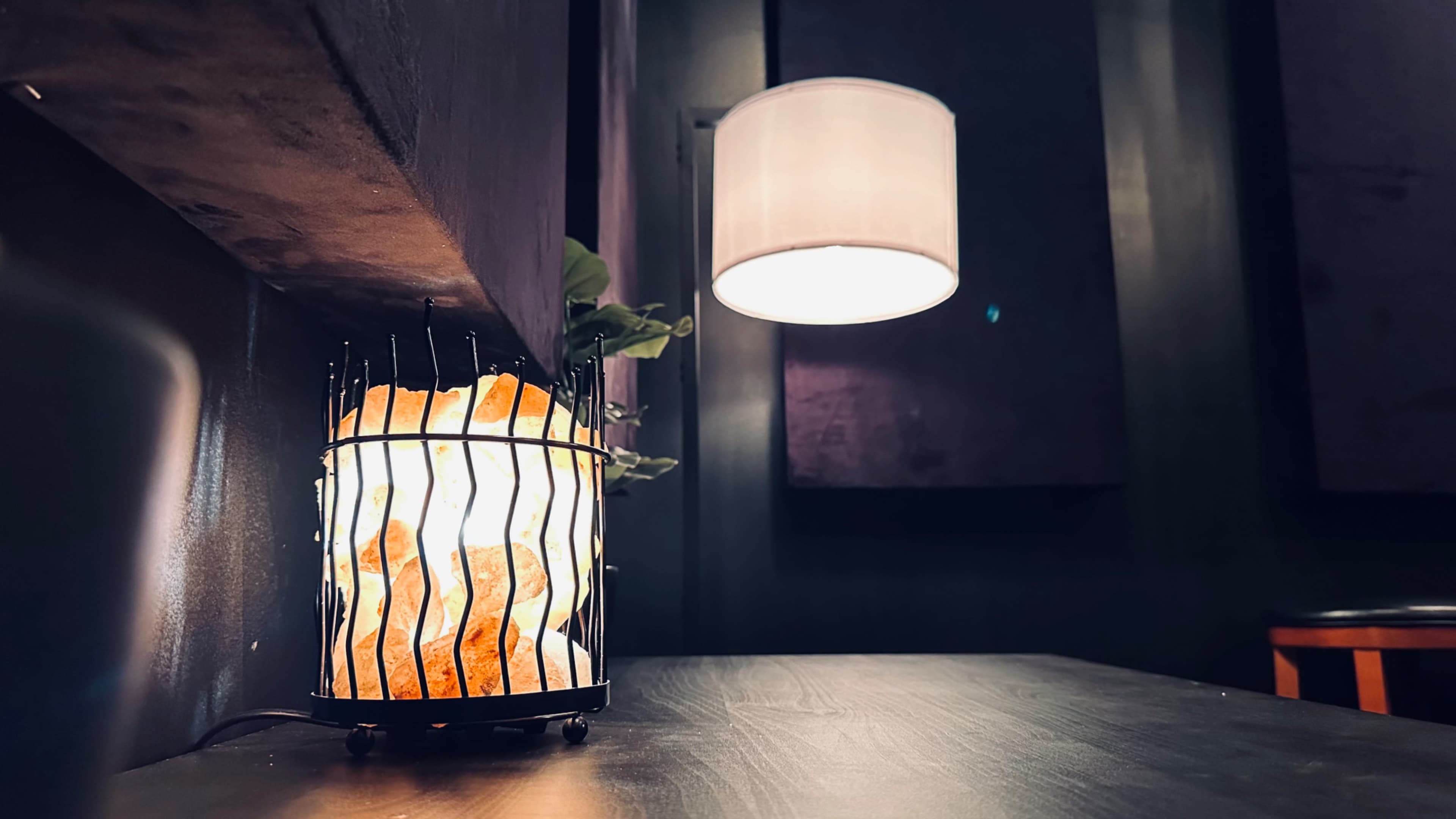 A wire basket filled with illuminated rocks sits on a dark wooden table beside a hanging lamp against a backdrop of dark walls.