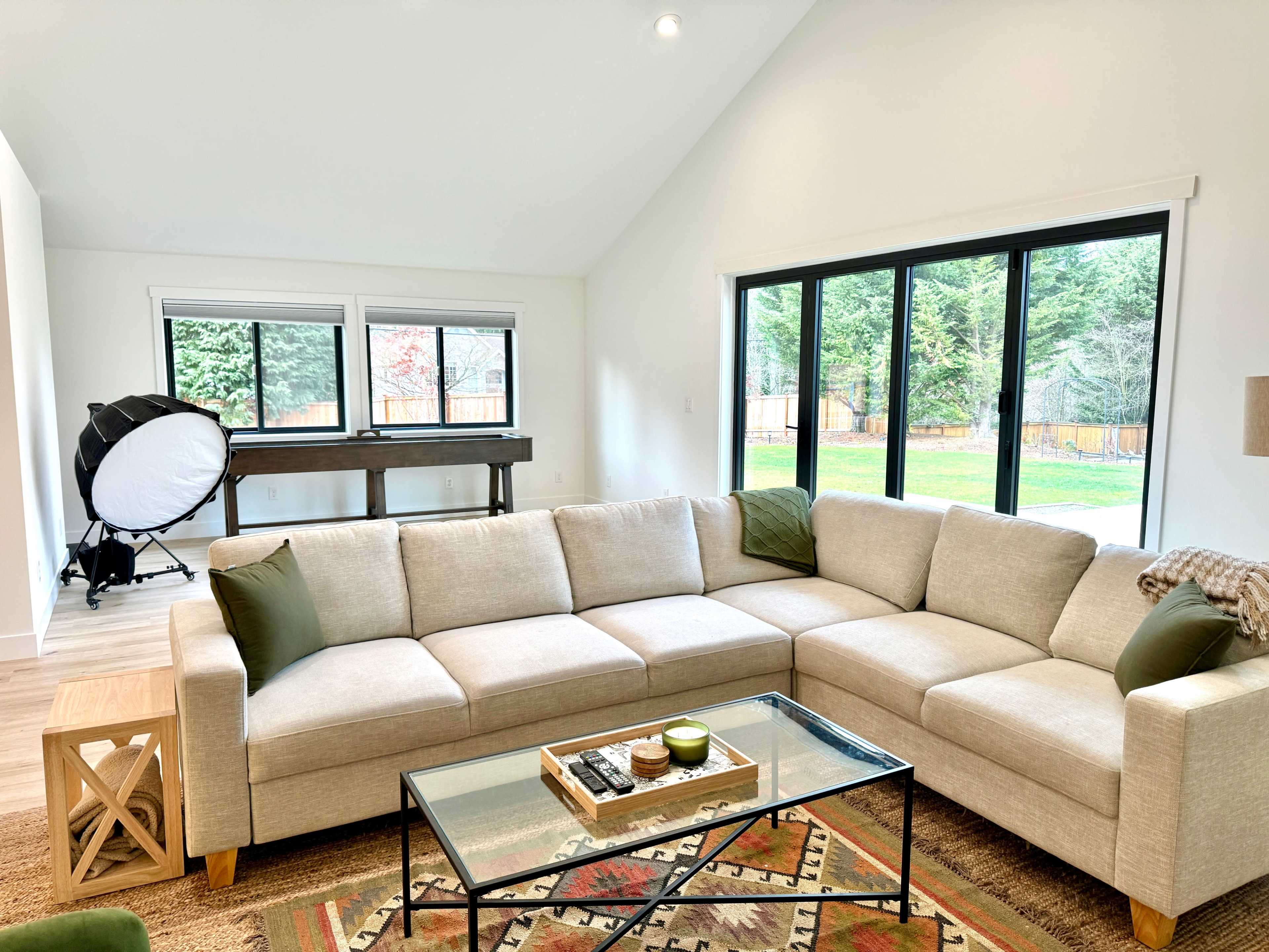 The image shows a modern living room with a beige sectional sofa, a glass coffee table, and large windows overlooking a green backyard.