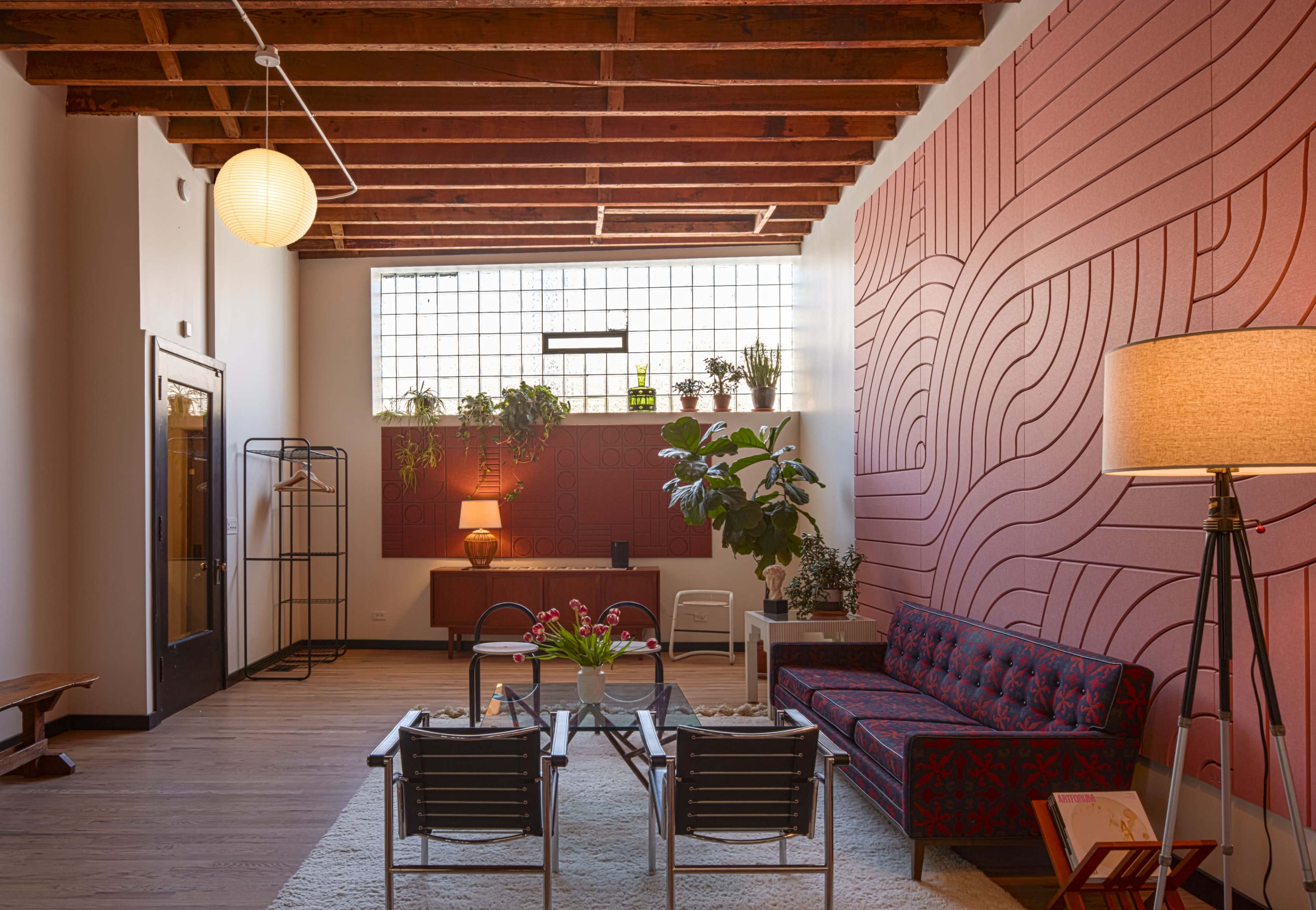 A modern living room featuring a patterned accent wall, plants on shelves, a combination of vintage and contemporary furniture, and wooden ceiling beams.