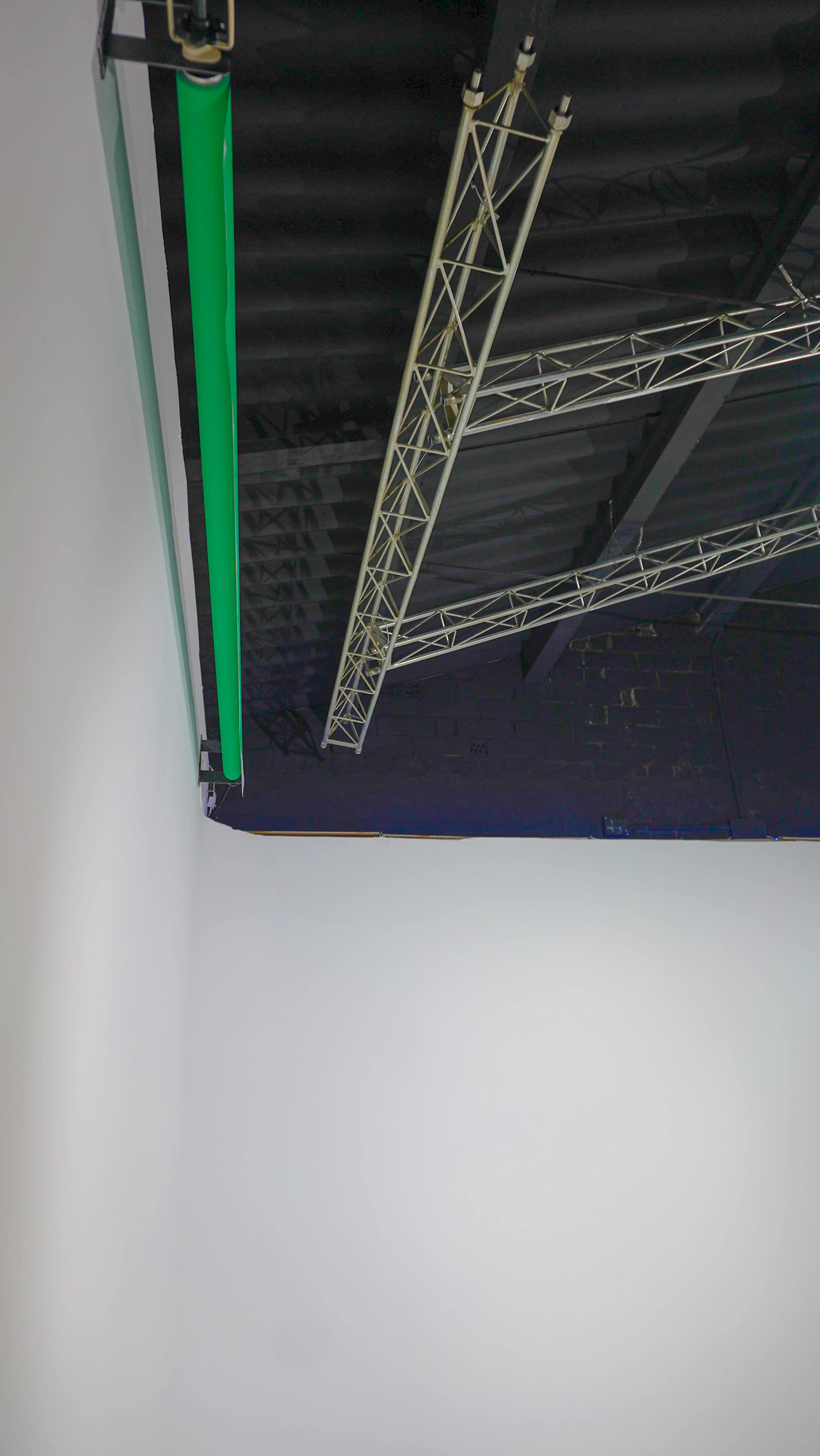 The image shows the corner of a room with a white wall and a ceiling featuring a green light and exposed truss support beams.