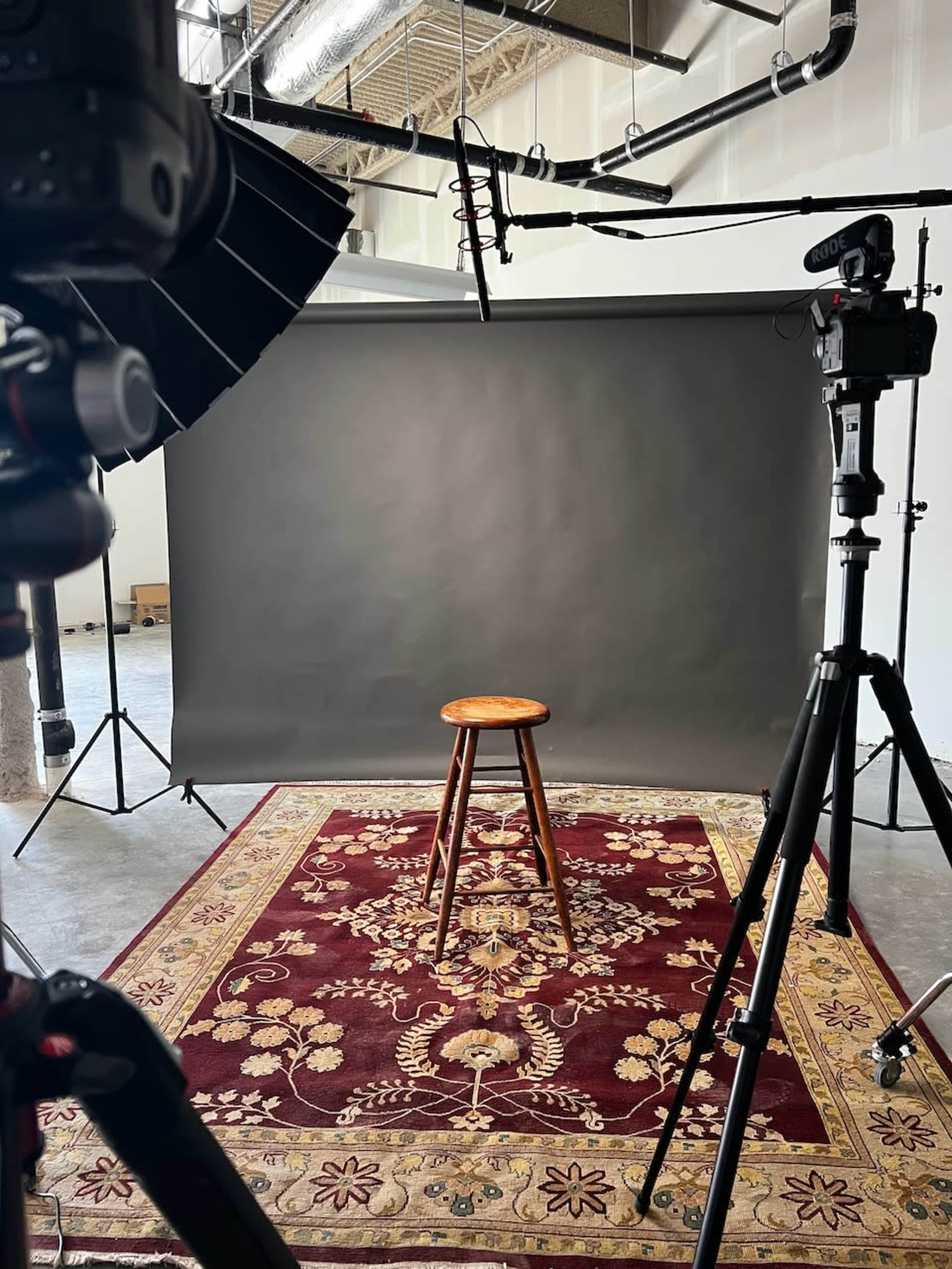 The image shows a photography studio setup with a wooden stool placed on a patterned rug in front of a gray backdrop, surrounded by camera equipment and lighting.
