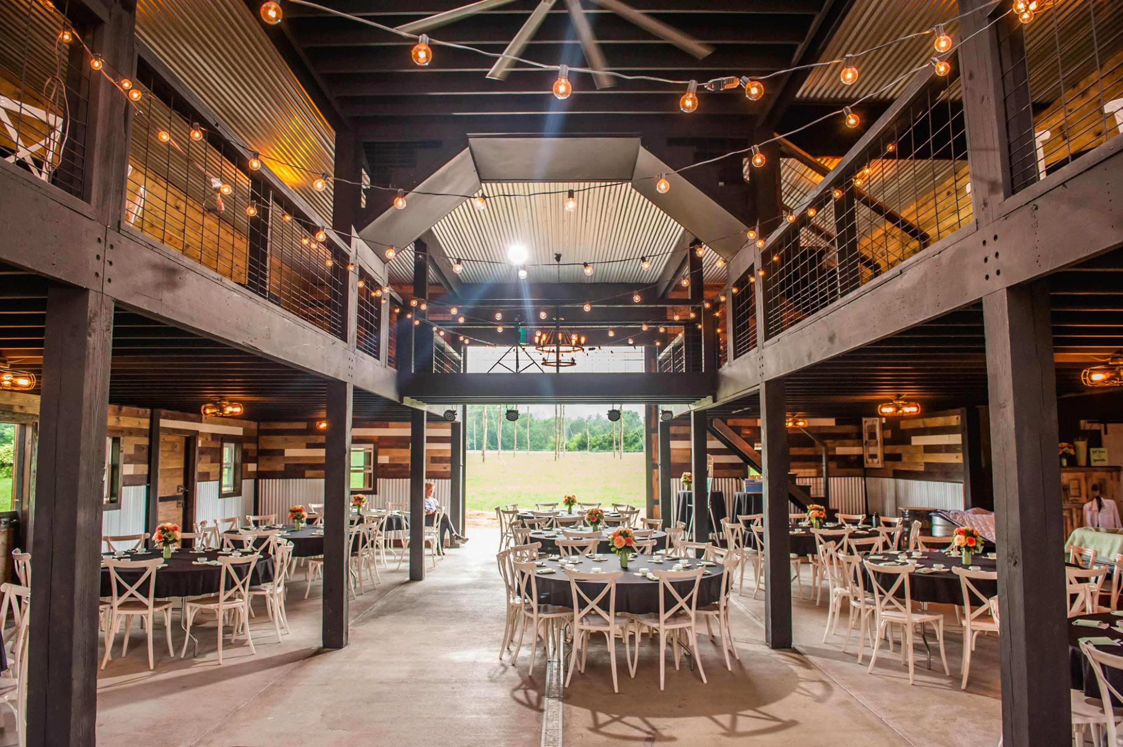 A spacious indoor venue set up for an event, featuring round tables with white chairs and string lights overhead.