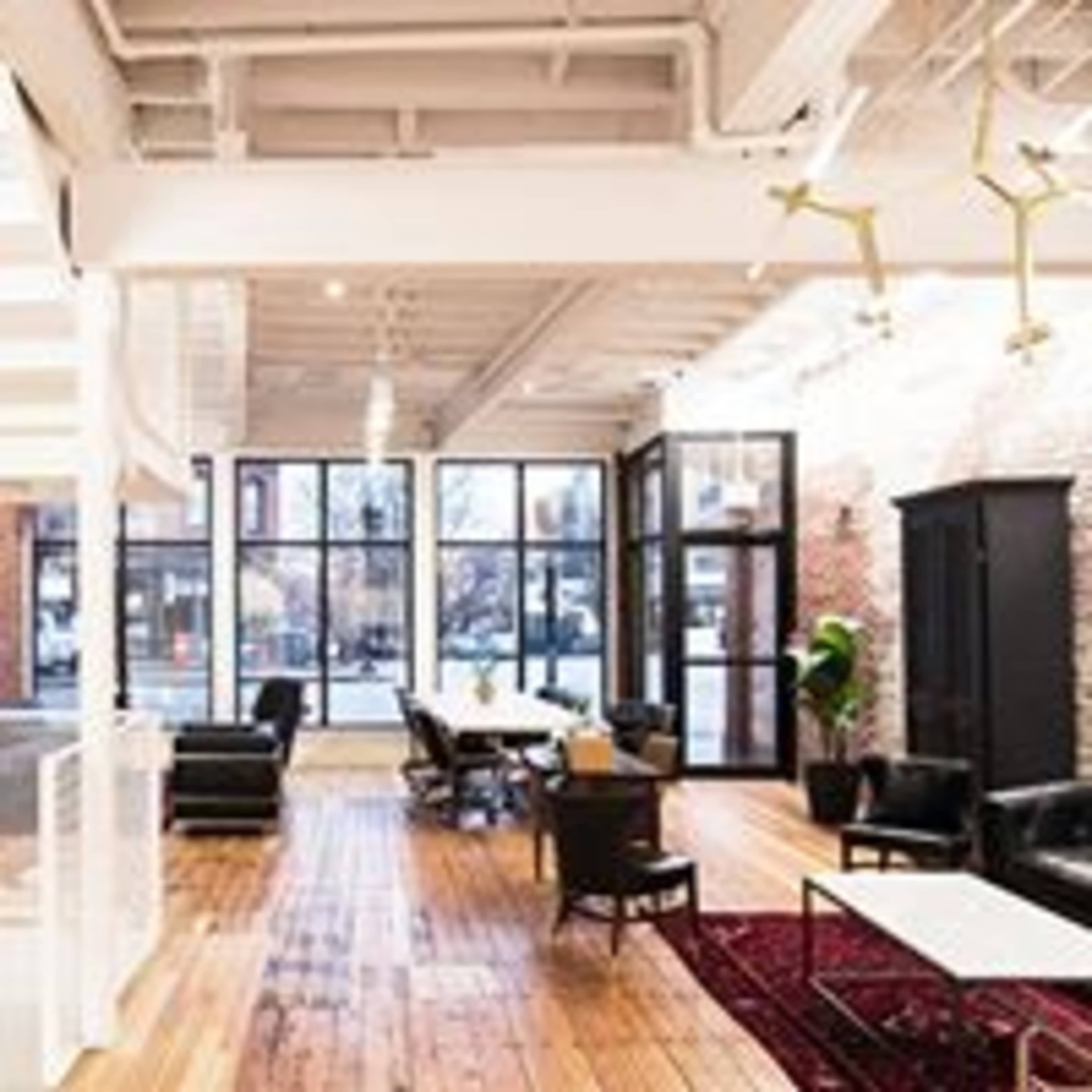 The image shows a modern, spacious office interior featuring wooden floors, large windows, and a mix of seating arrangements, including black leather chairs and a table.