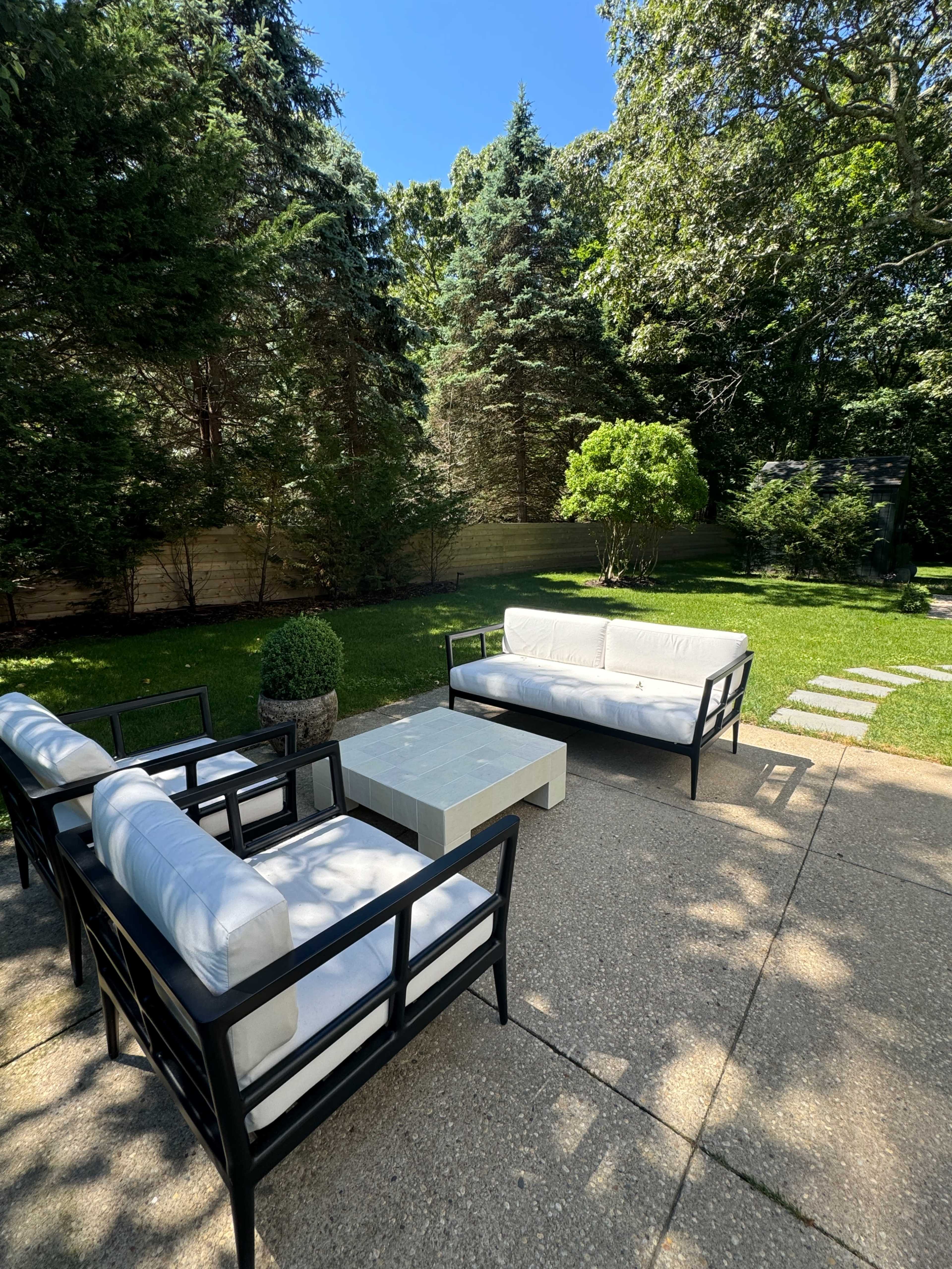 A patio area features four black-framed chairs with white cushions and a stone coffee table, surrounded by grass and trees.