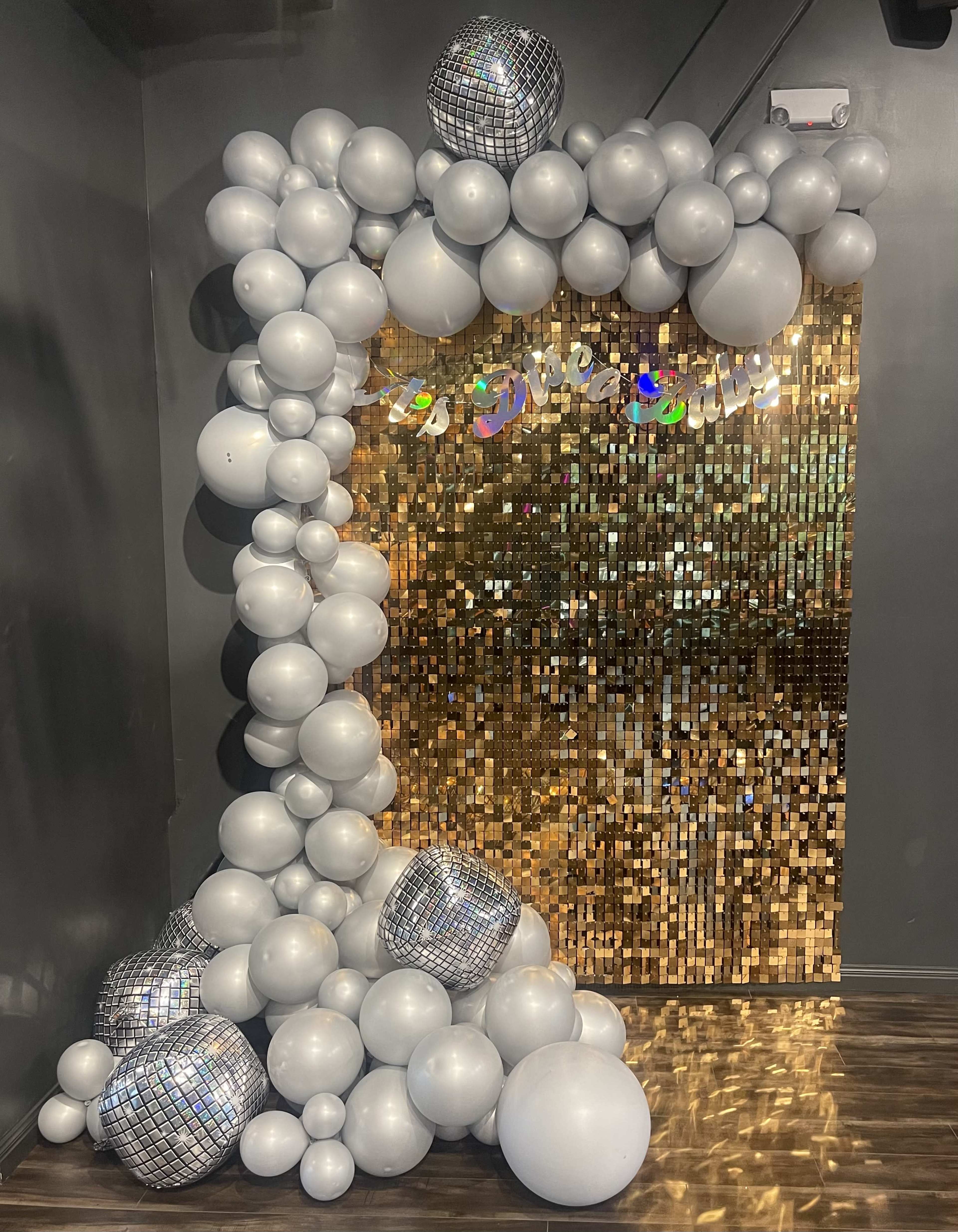 A decorated entrance featuring a large arch made of silver balloons alongside a shiny gold sequin backdrop.