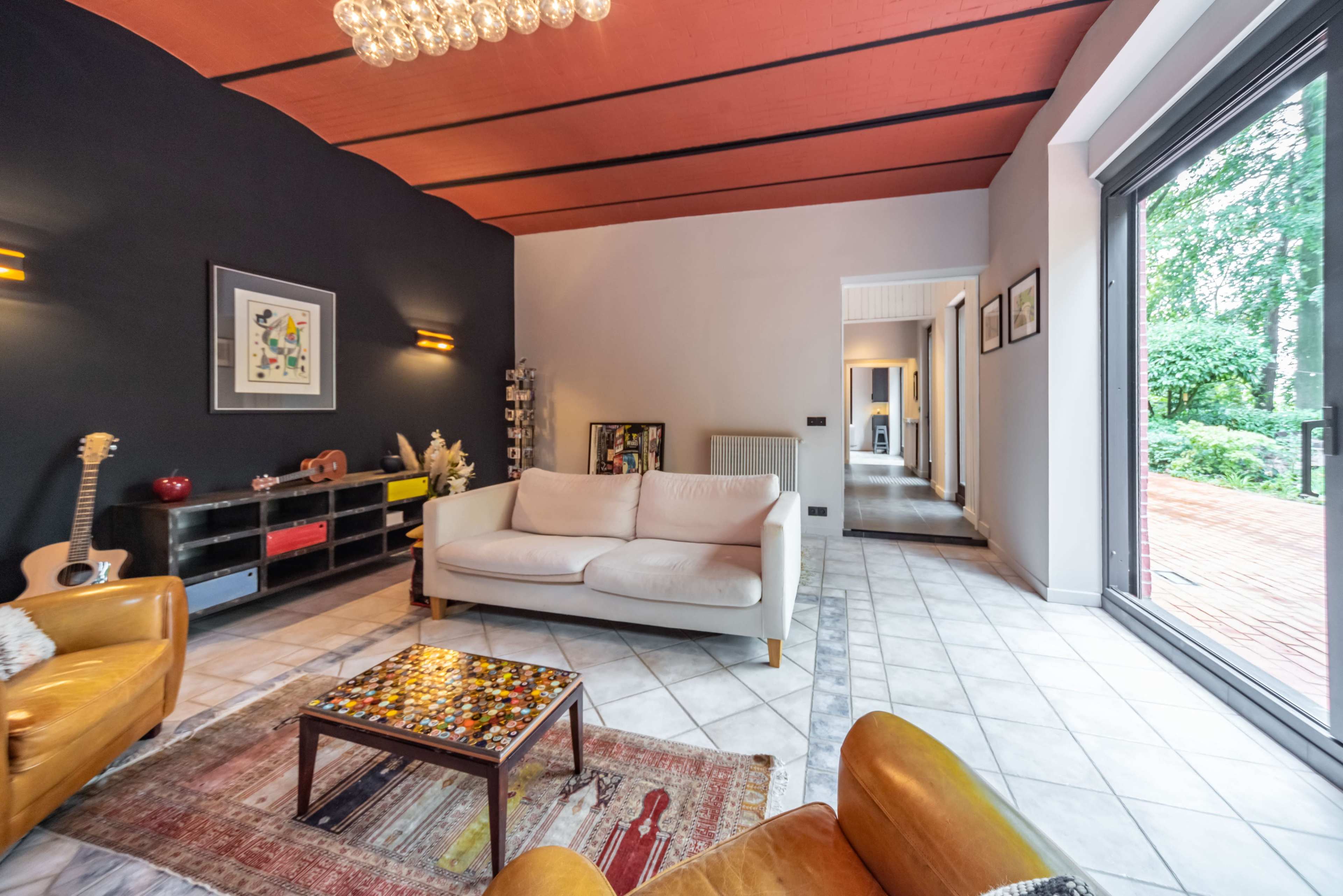 A modern living room features a white sofa, two leather chairs, a colorful coffee table, and large windows overlooking a garden.