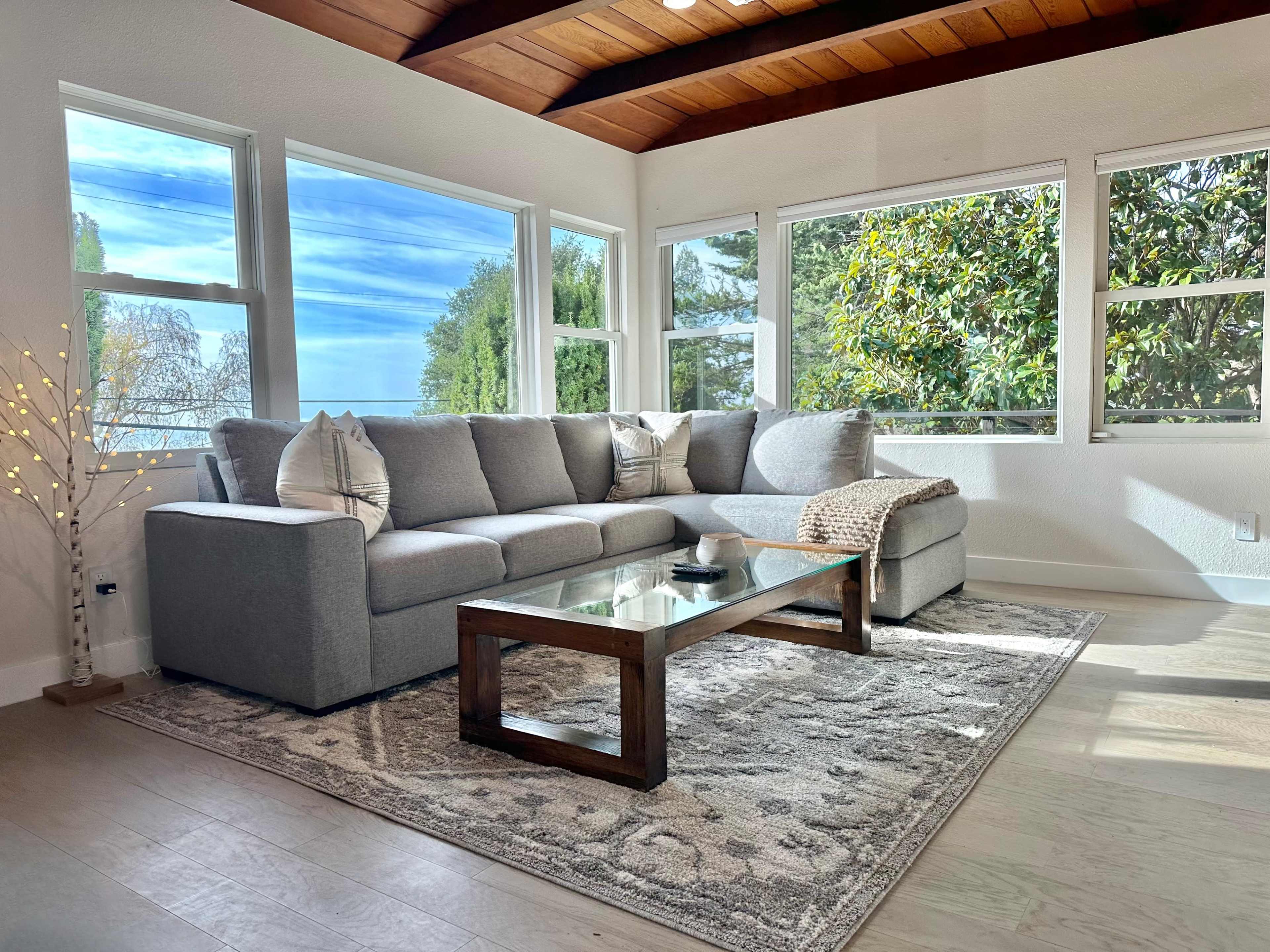 A modern living room features a gray sectional sofa, a glass coffee table, and large windows allowing natural light to fill the space.