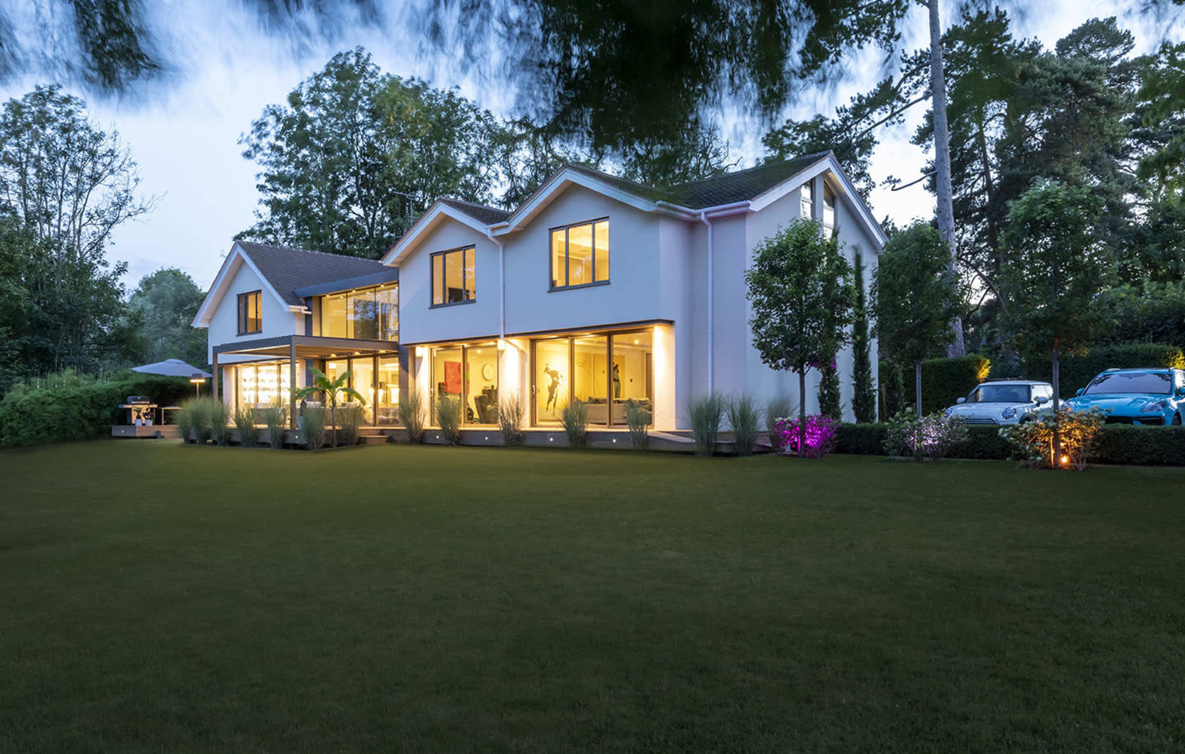 A modern two-story house with large windows is surrounded by a well-maintained lawn and gardens in the evening light.