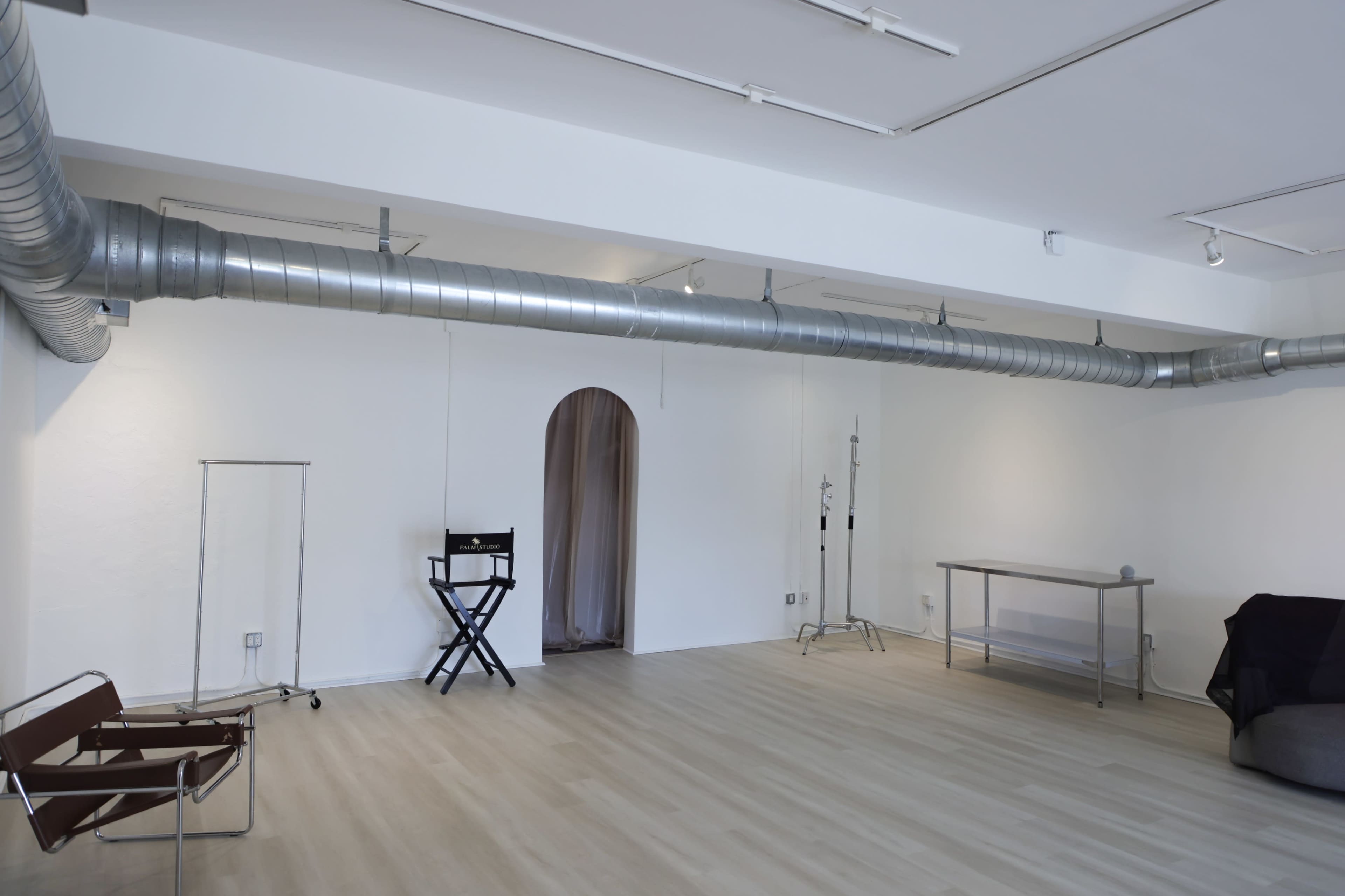 The image shows an empty, modern studio space with wooden flooring, white walls, and a prominent metal duct running along the ceiling.