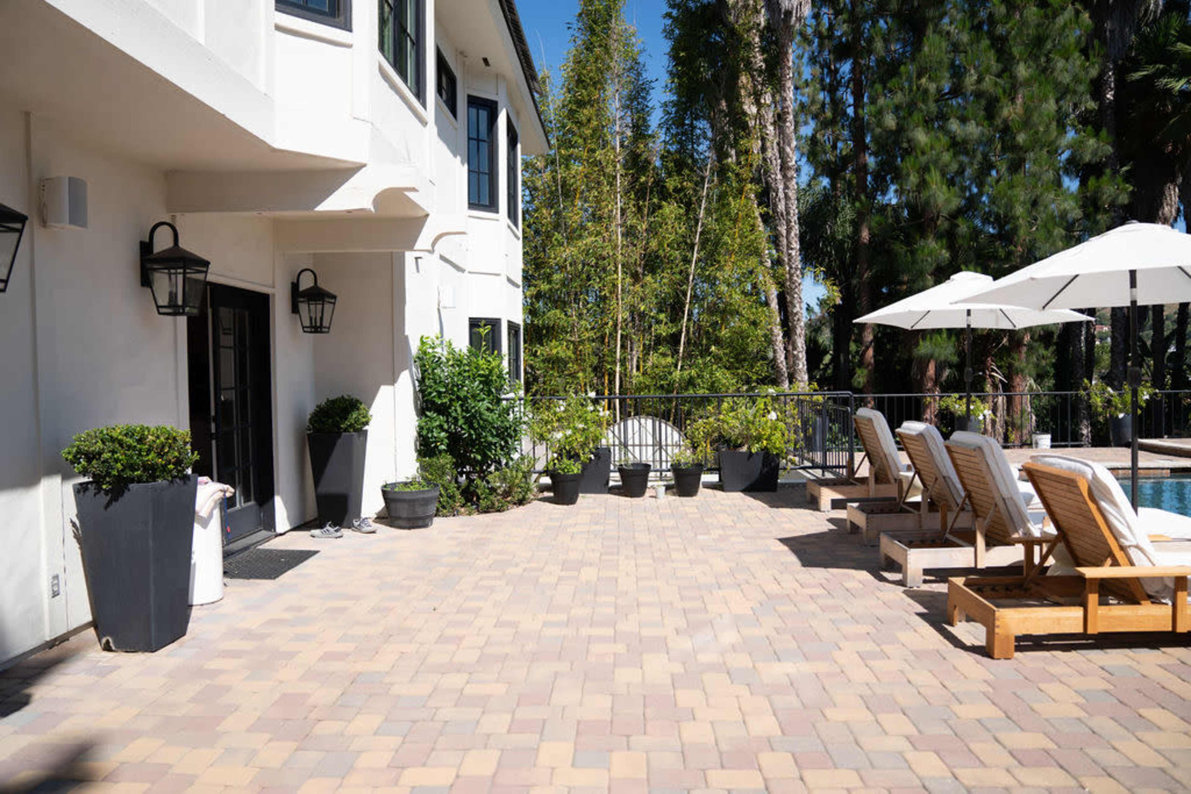 A patio area with lounge chairs and umbrellas beside a swimming pool, surrounded by greenery and a modern white house.