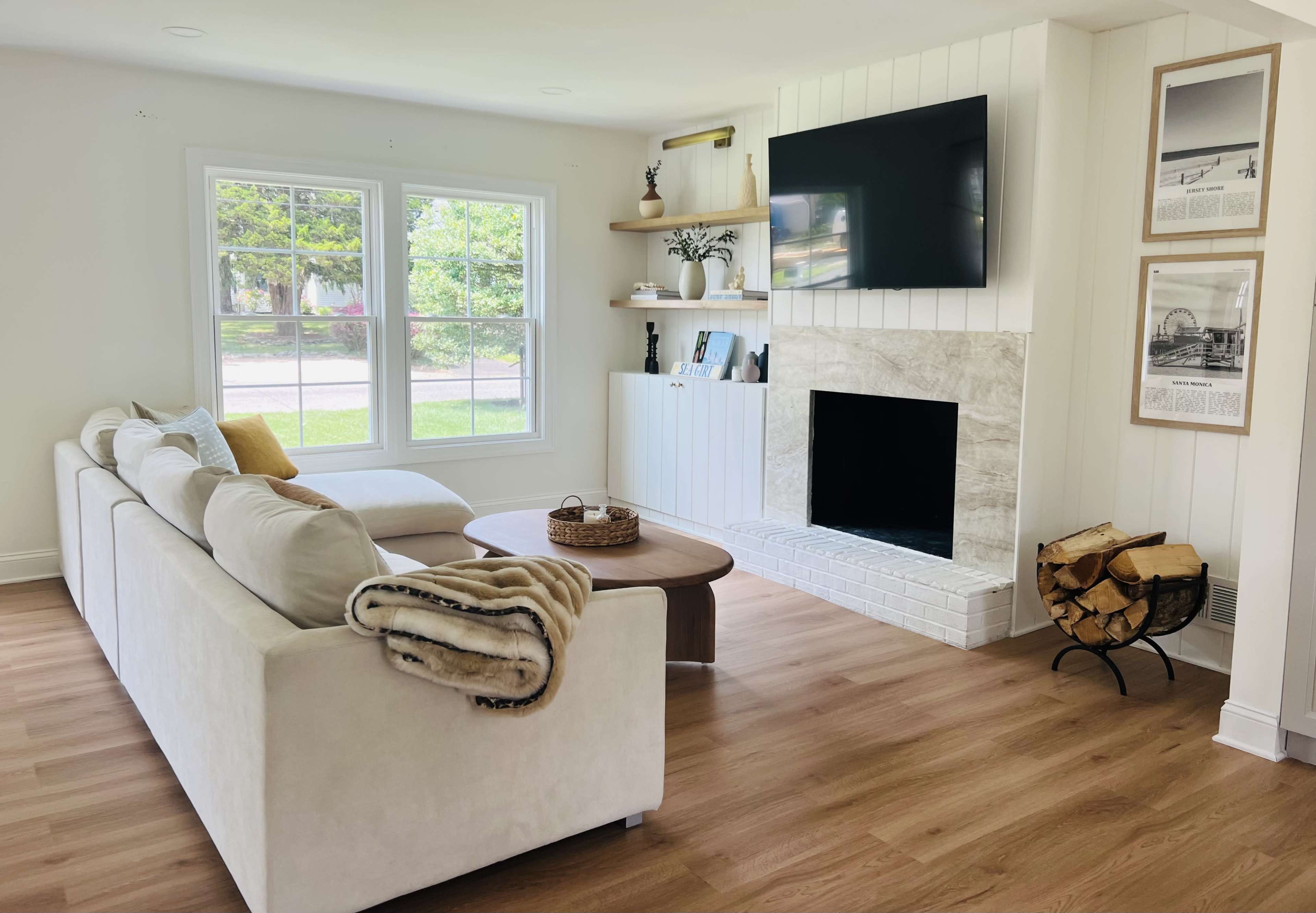 A bright and modern living room featuring a light-colored sofa, a round coffee table, a wall-mounted TV, and a fireplace with stacked firewood beside it.