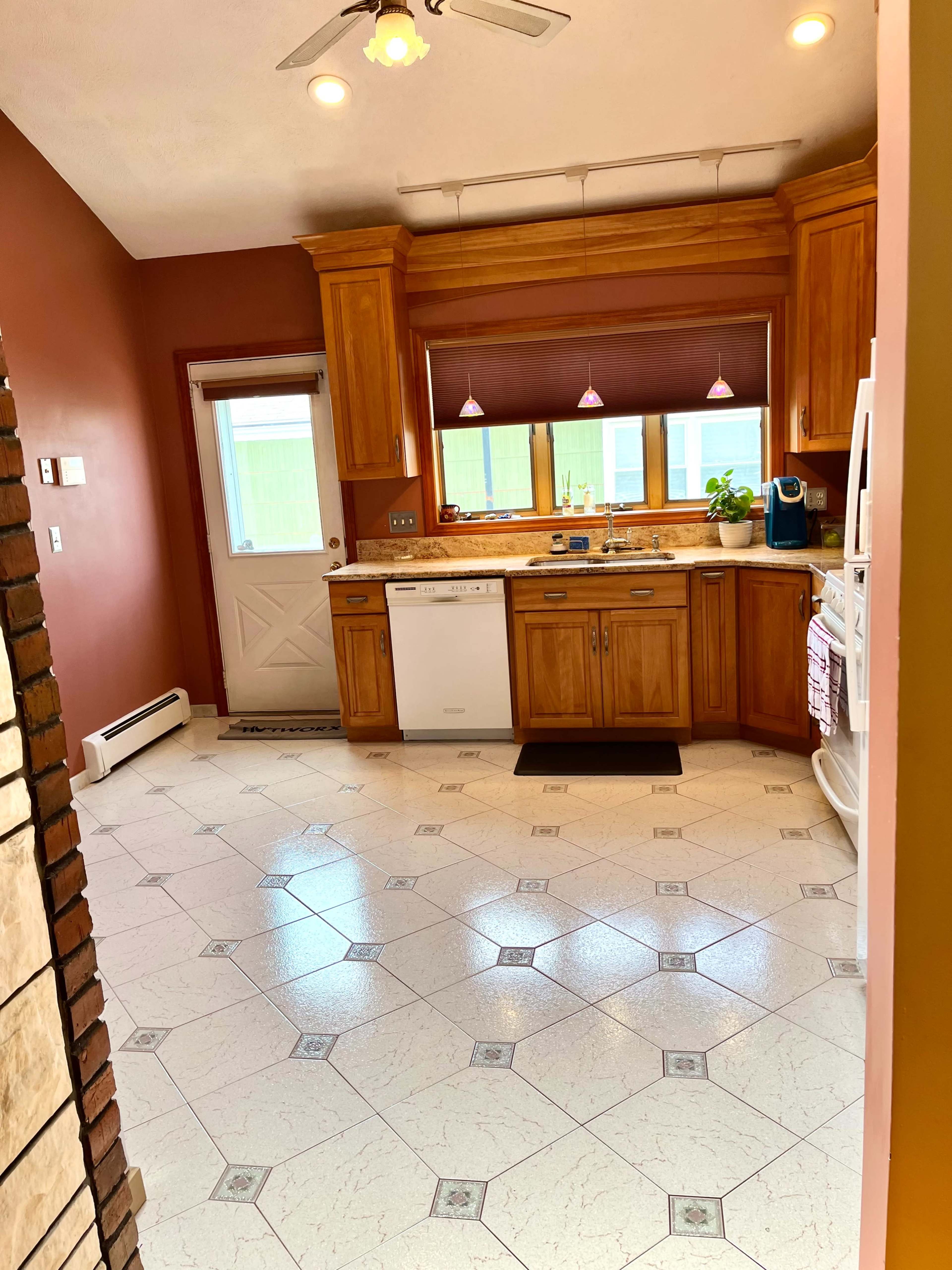 A kitchen features wooden cabinetry, a dishwasher, and a door leading outside, with tiled flooring and overhead lighting.