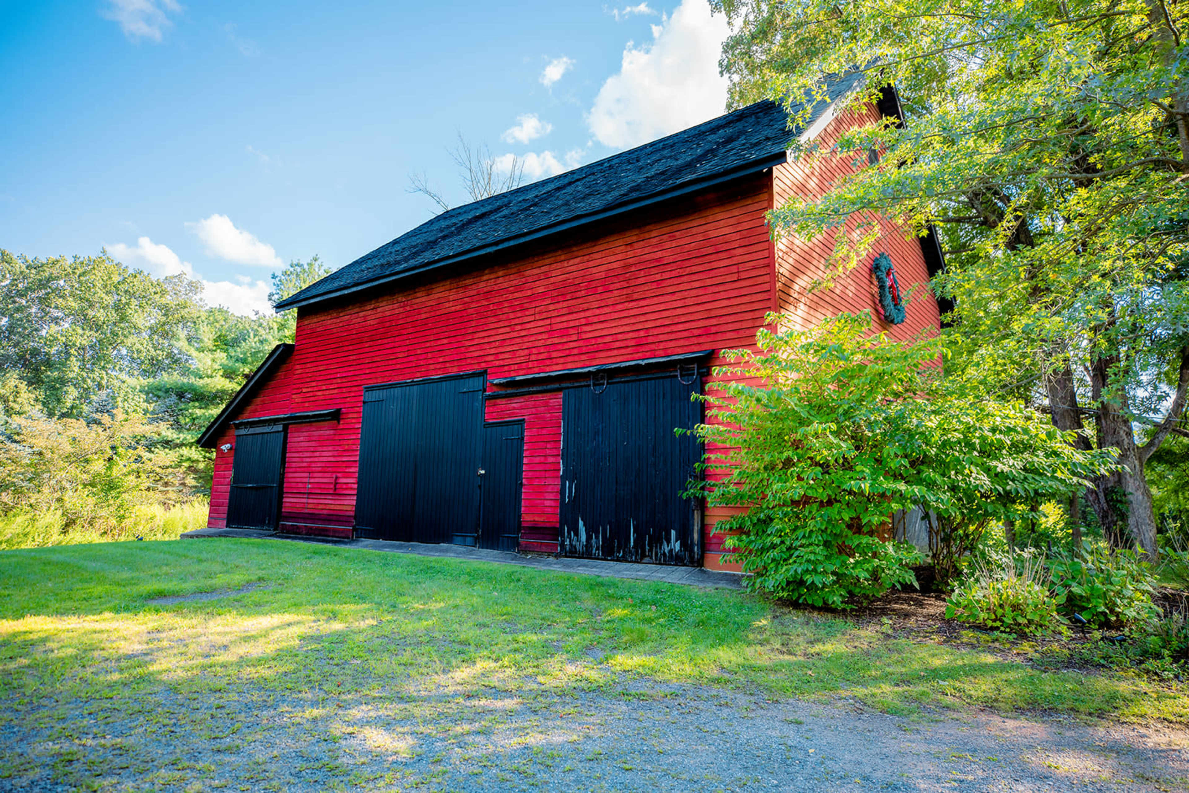 A red barn with black doors stands next to a patch of green grass and trees against a clear blue sky.