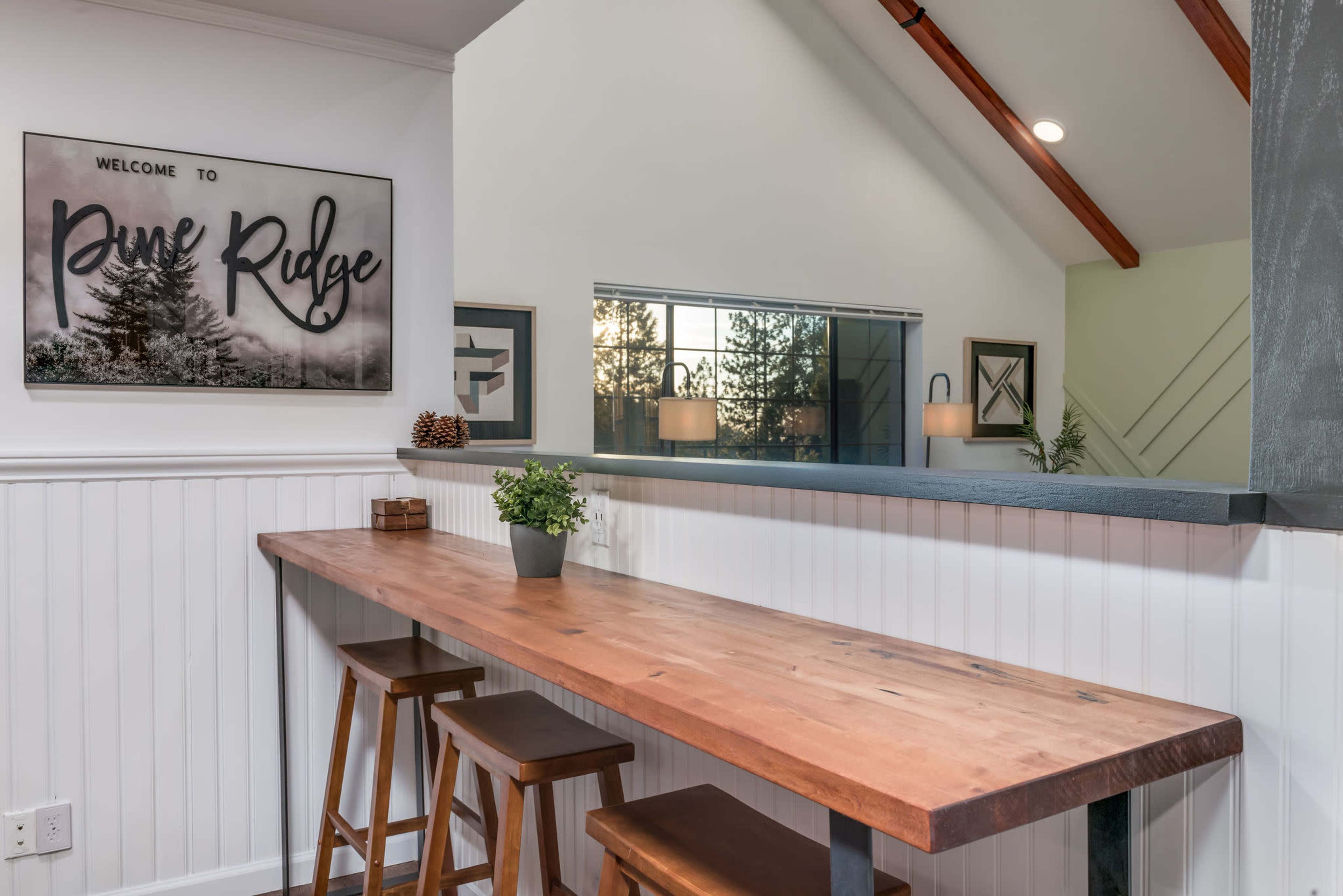 A wooden bar table with three stools sits against a white paneled wall, featuring a wall art piece that reads "Welcome to Pine Ridge."