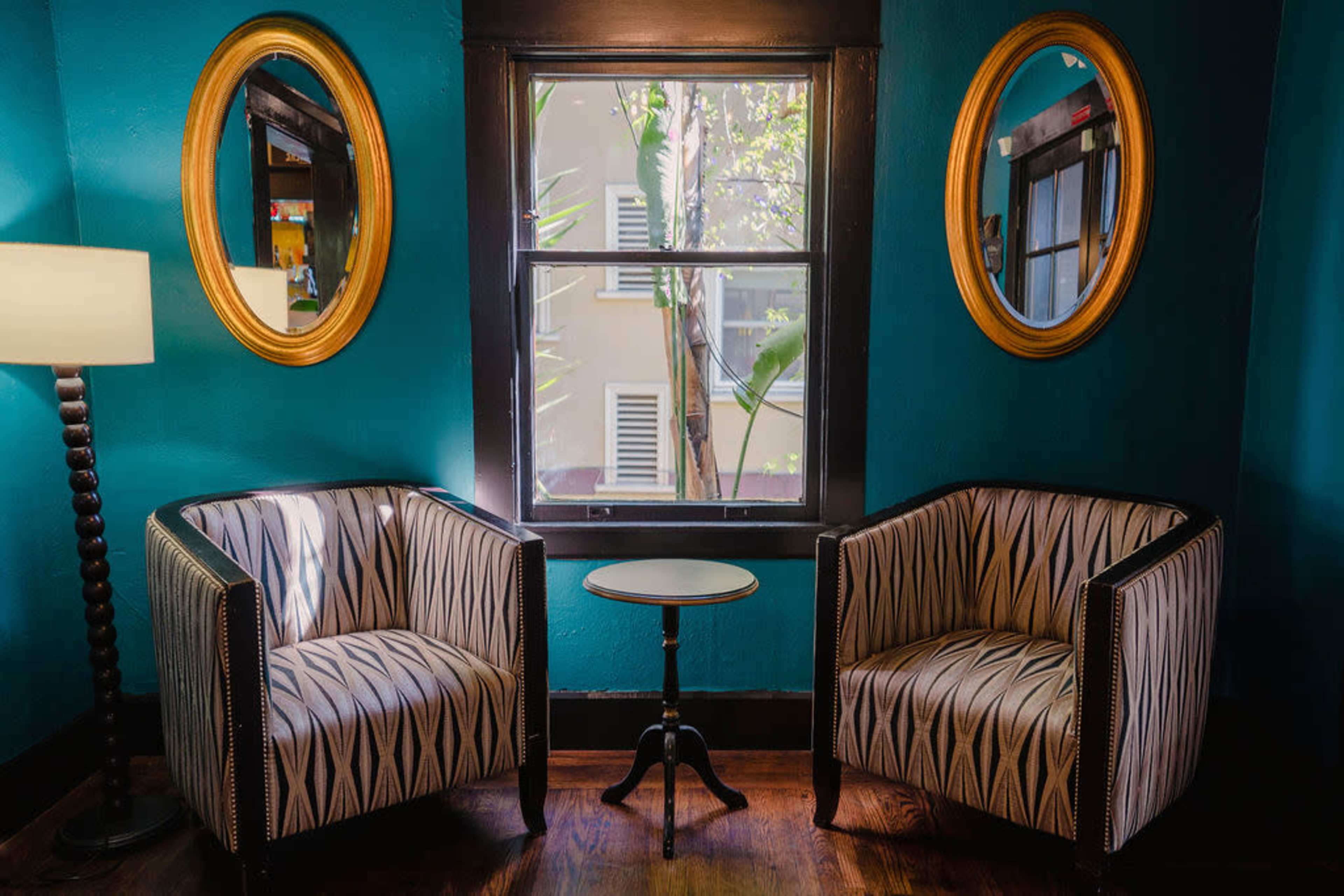 Two patterned armchairs face each other with a small round table in between, set against a teal wall with a window featuring round mirrors on either side.