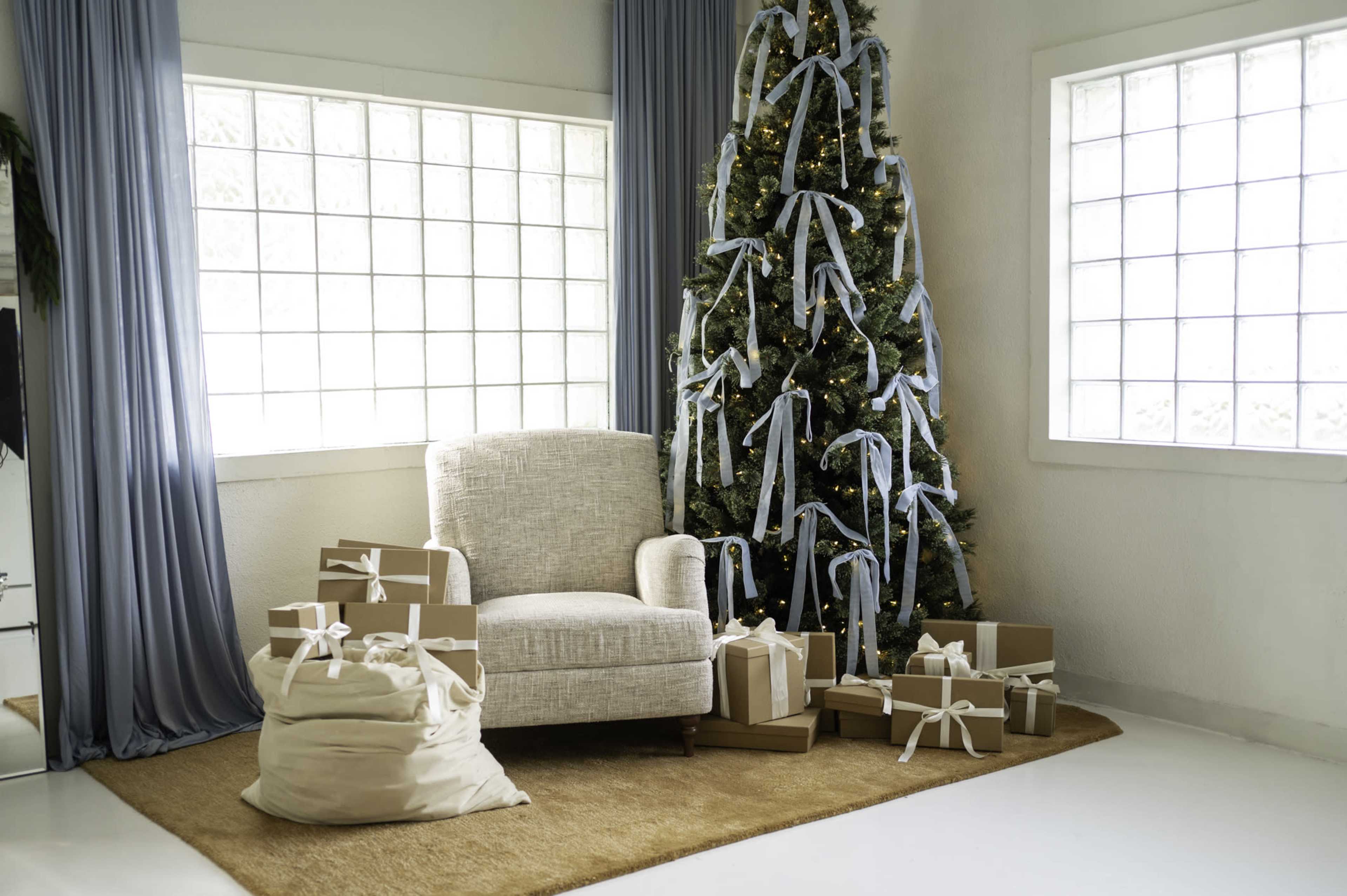 A neatly decorated Christmas tree adorned with blue ribbons stands beside a cozy chair and an assortment of wrapped gifts on a beige rug.