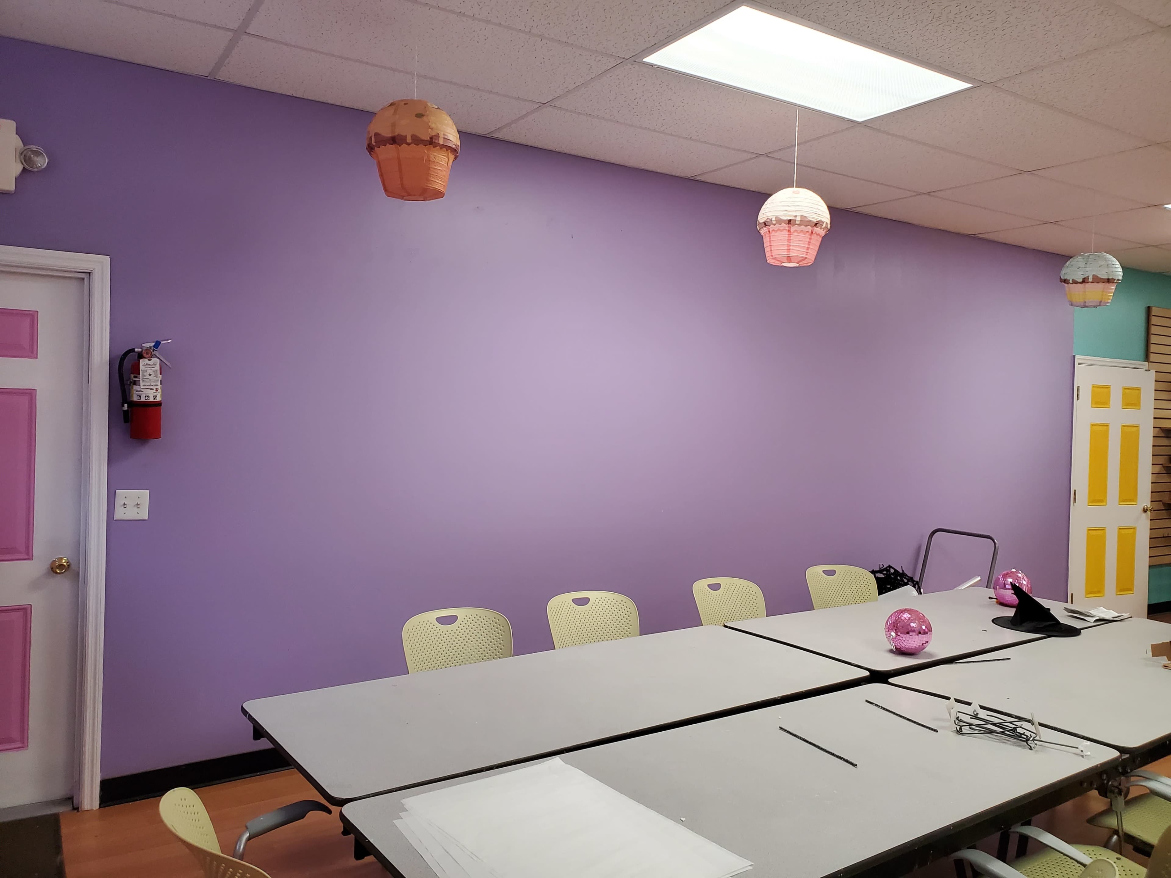 The image shows a light purple wall with a long table set up in front of it, surrounded by several chairs, and adorned with decorative hanging lanterns above.