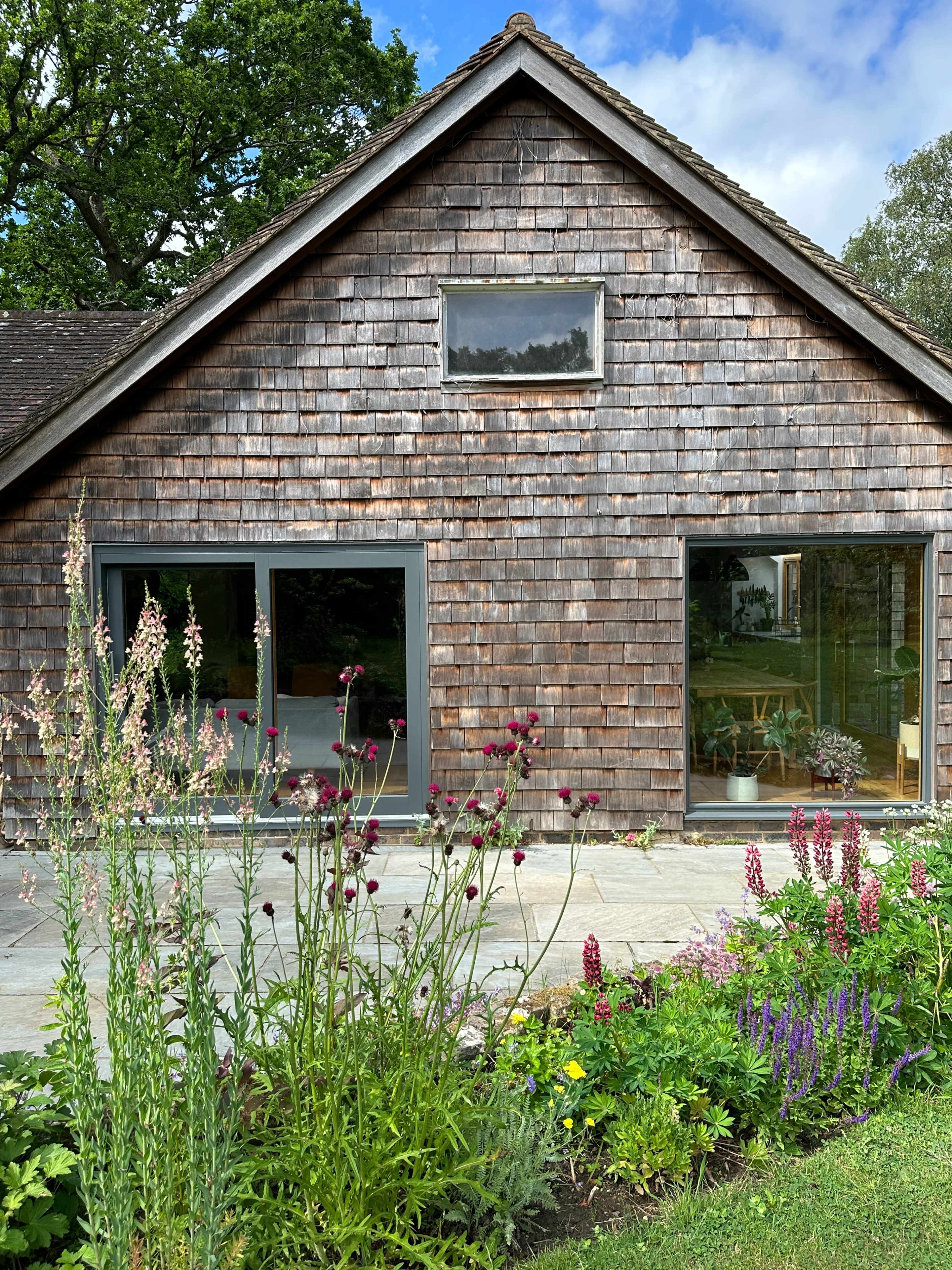 A wooden house with a shingled exterior features large windows, surrounded by a well-maintained garden with various flowering plants.