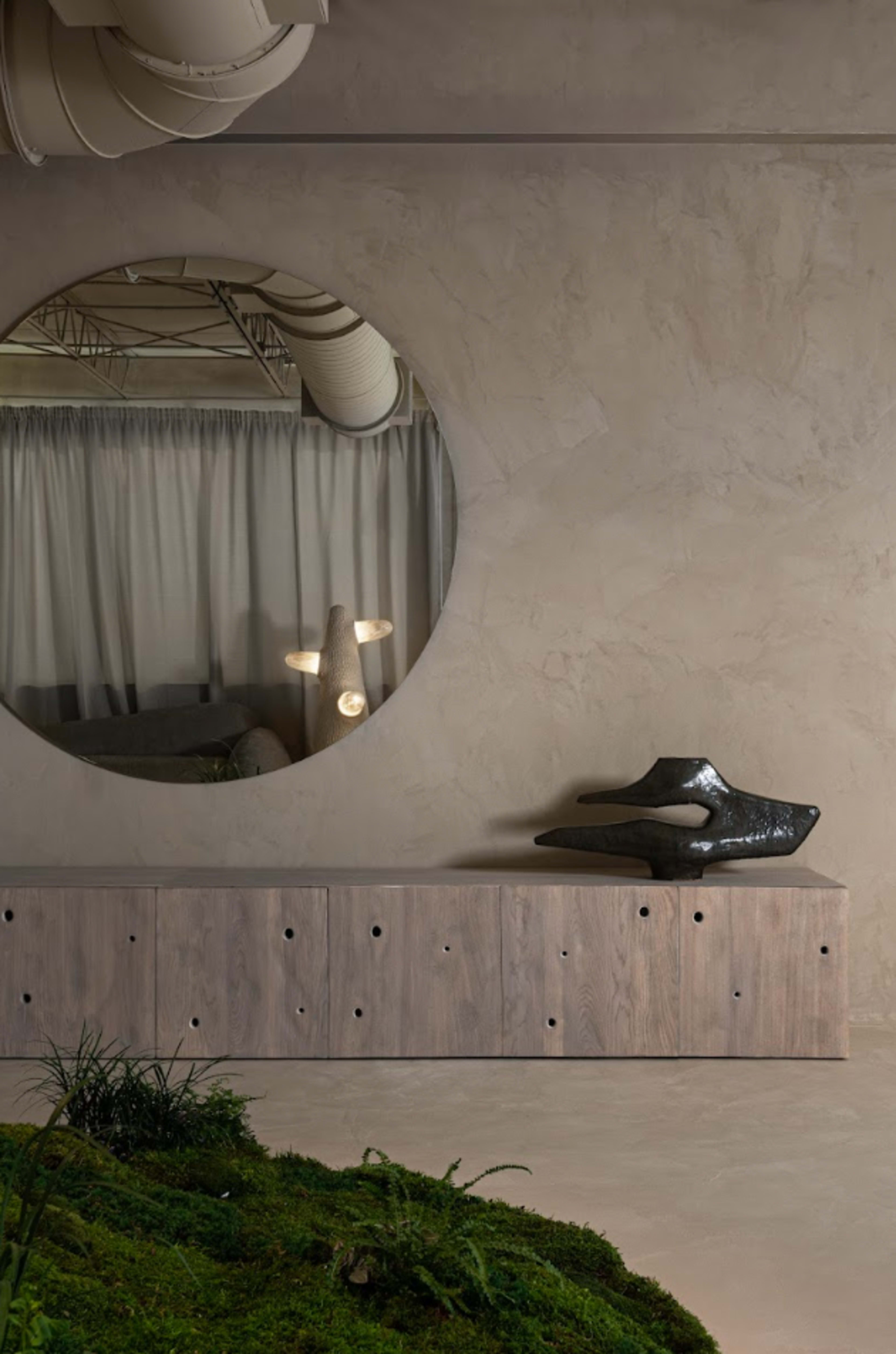 A circular mirror hangs on a textured wall above a wooden cabinet, which displays a sculptural piece alongside greenery on the floor.