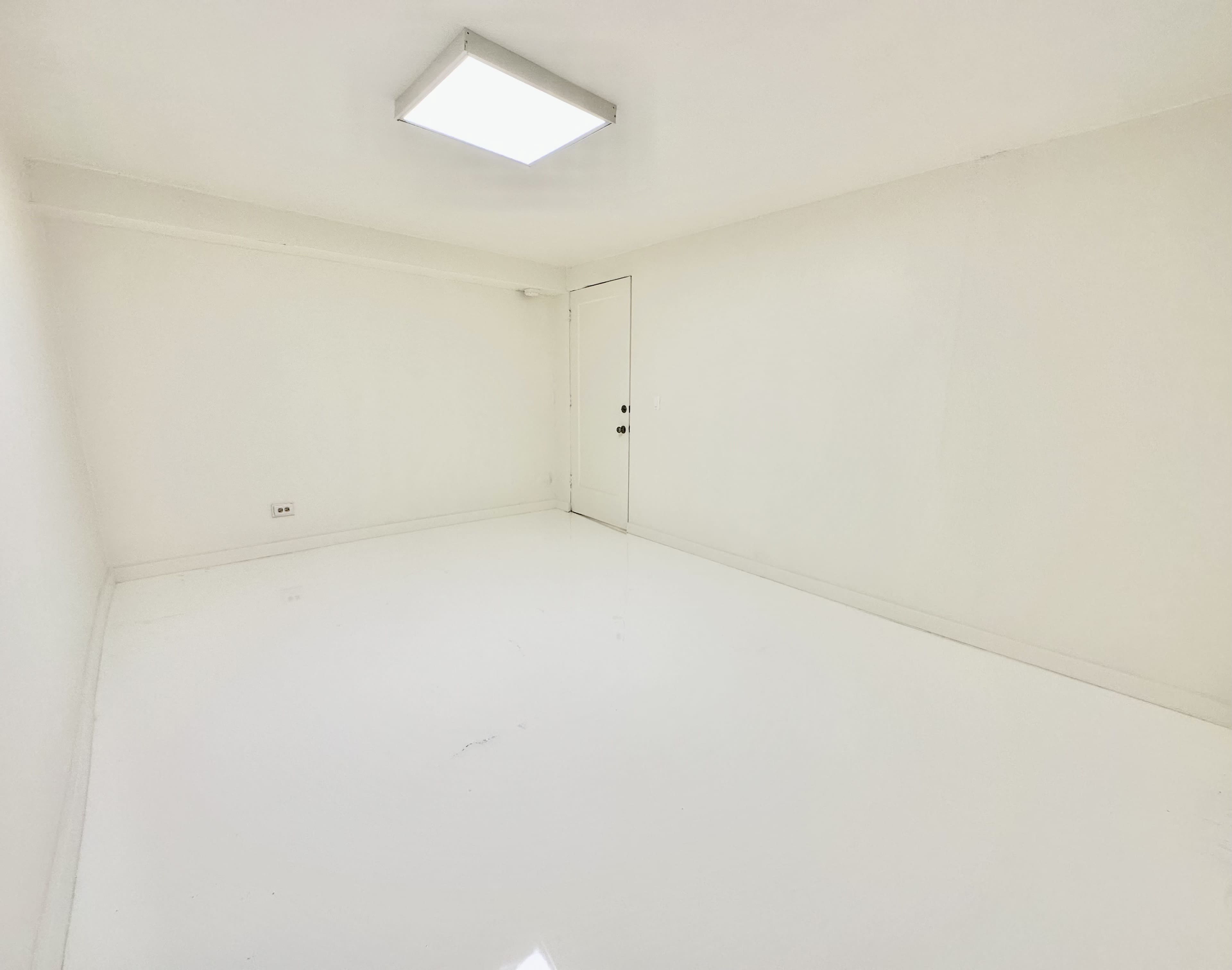 An empty room with white walls and a glossy white floor, illuminated by a ceiling light.