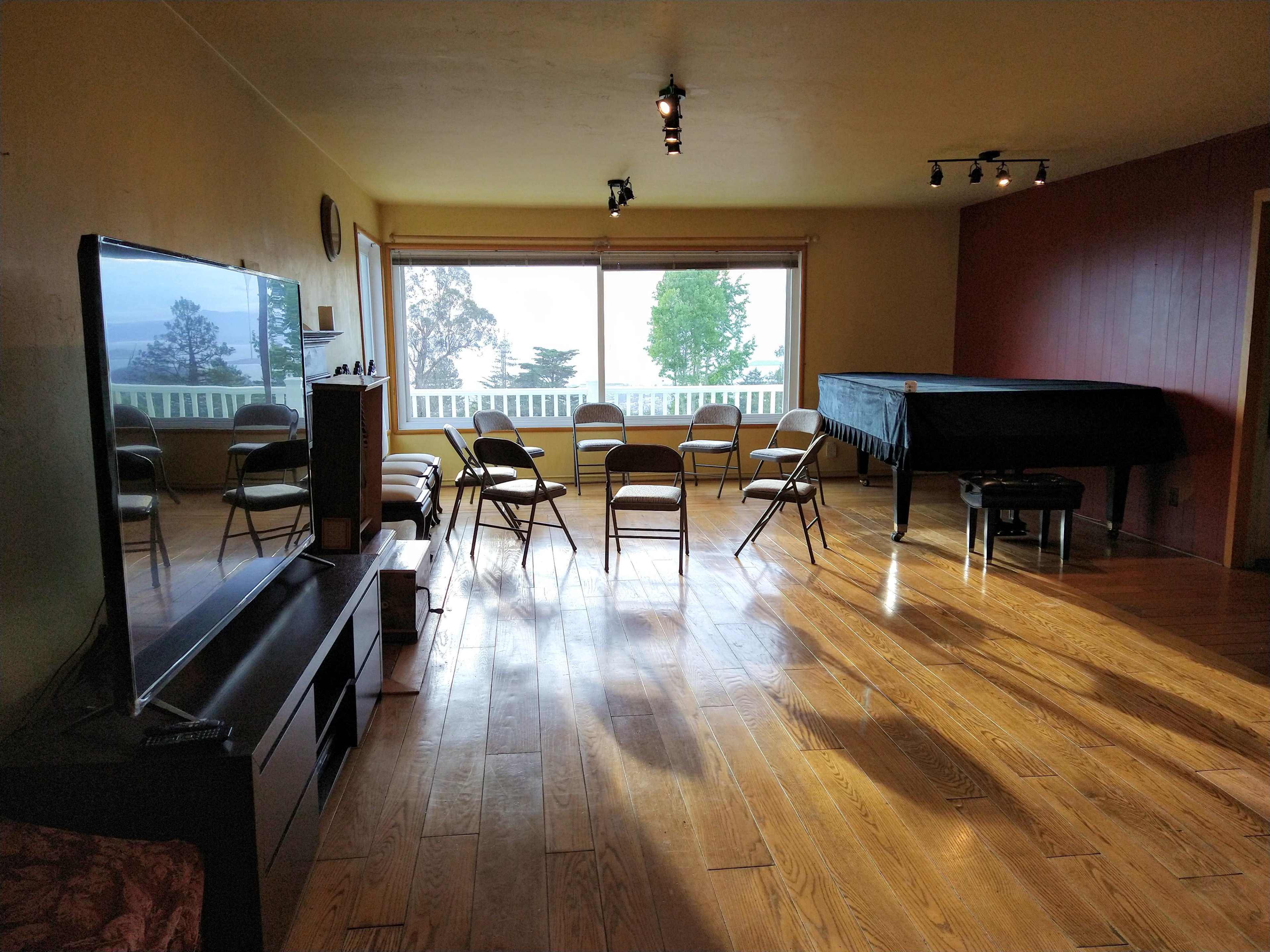 A large, empty room with wooden floors features a set of eight folding chairs arranged in a circle, a grand piano in the corner, and a large window that overlooks a scenic view.