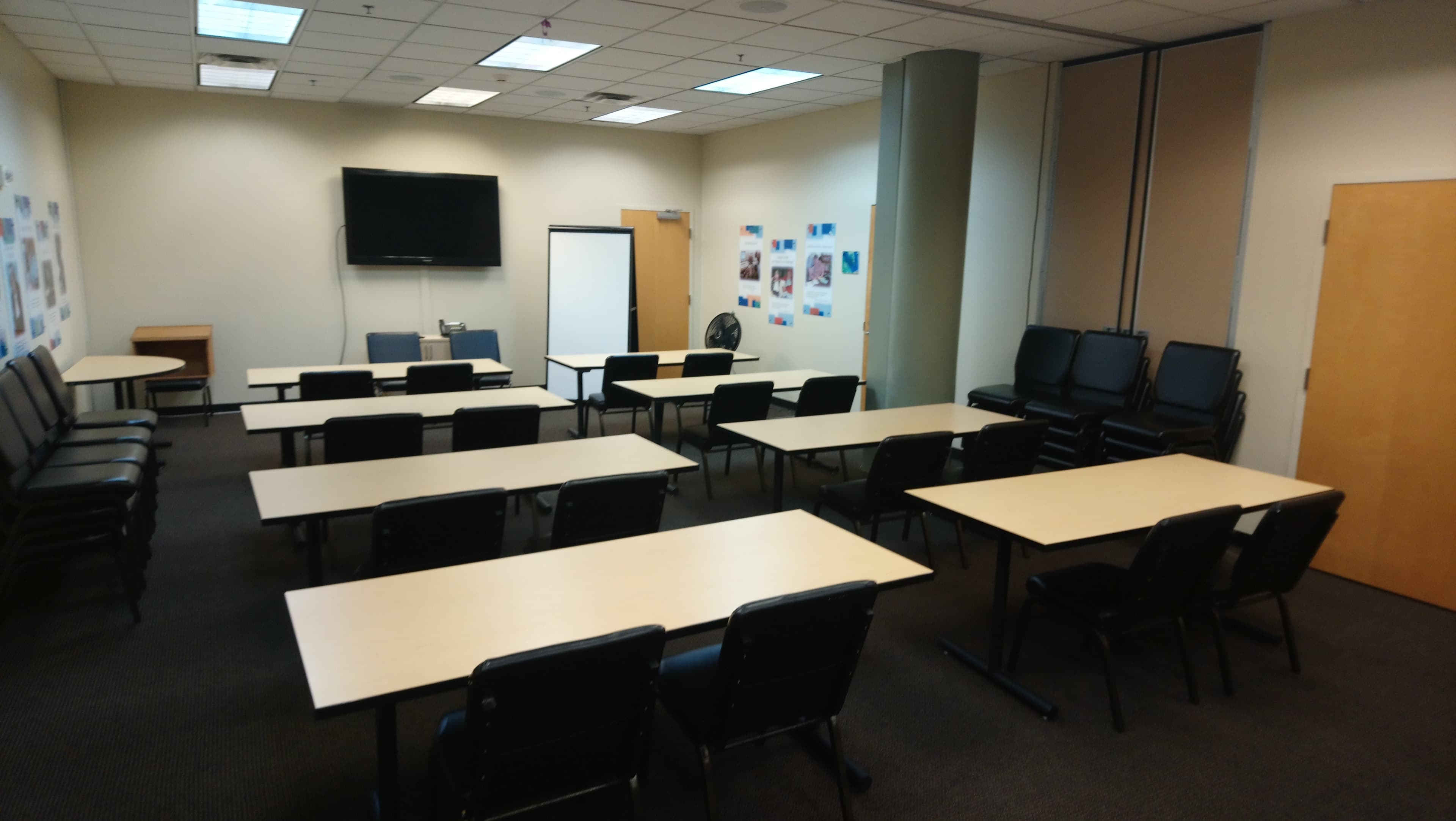 A training room set up with multiple rows of tables and chairs, a blank whiteboard, and a television on the wall.