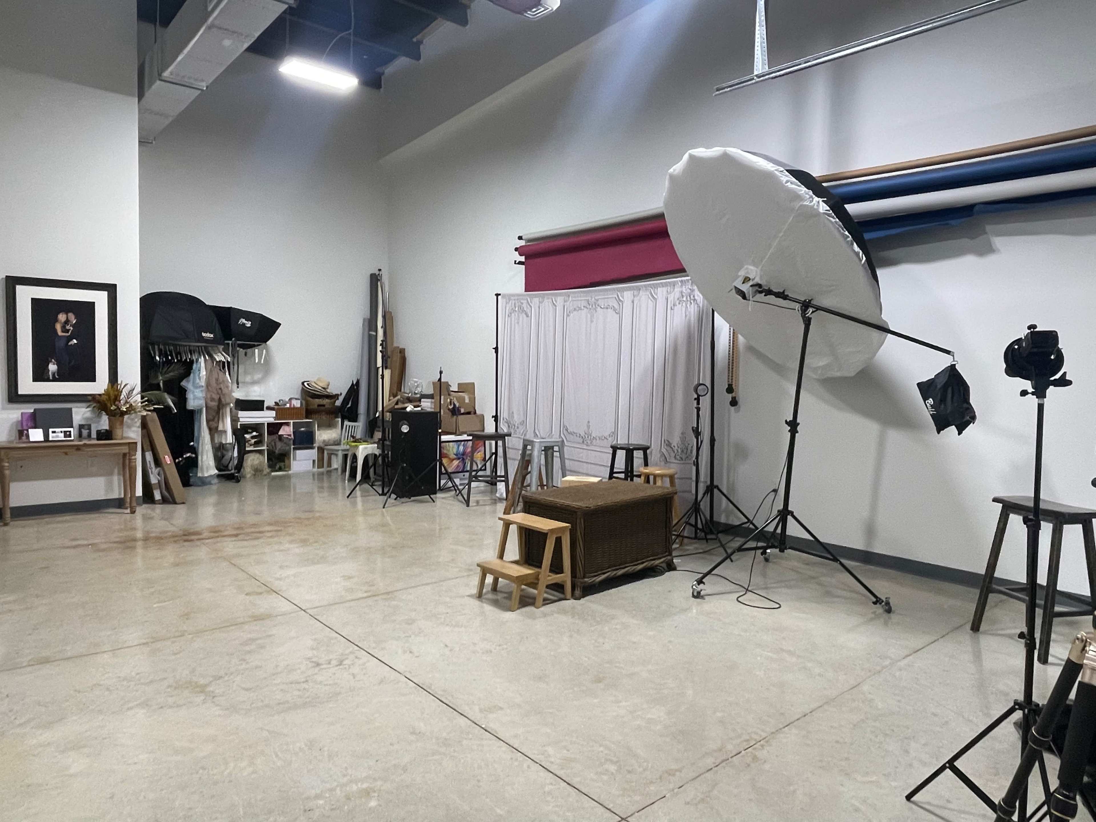 A photography studio contains various backdrops, lighting equipment, and props arranged around a spacious, uncluttered concrete floor.