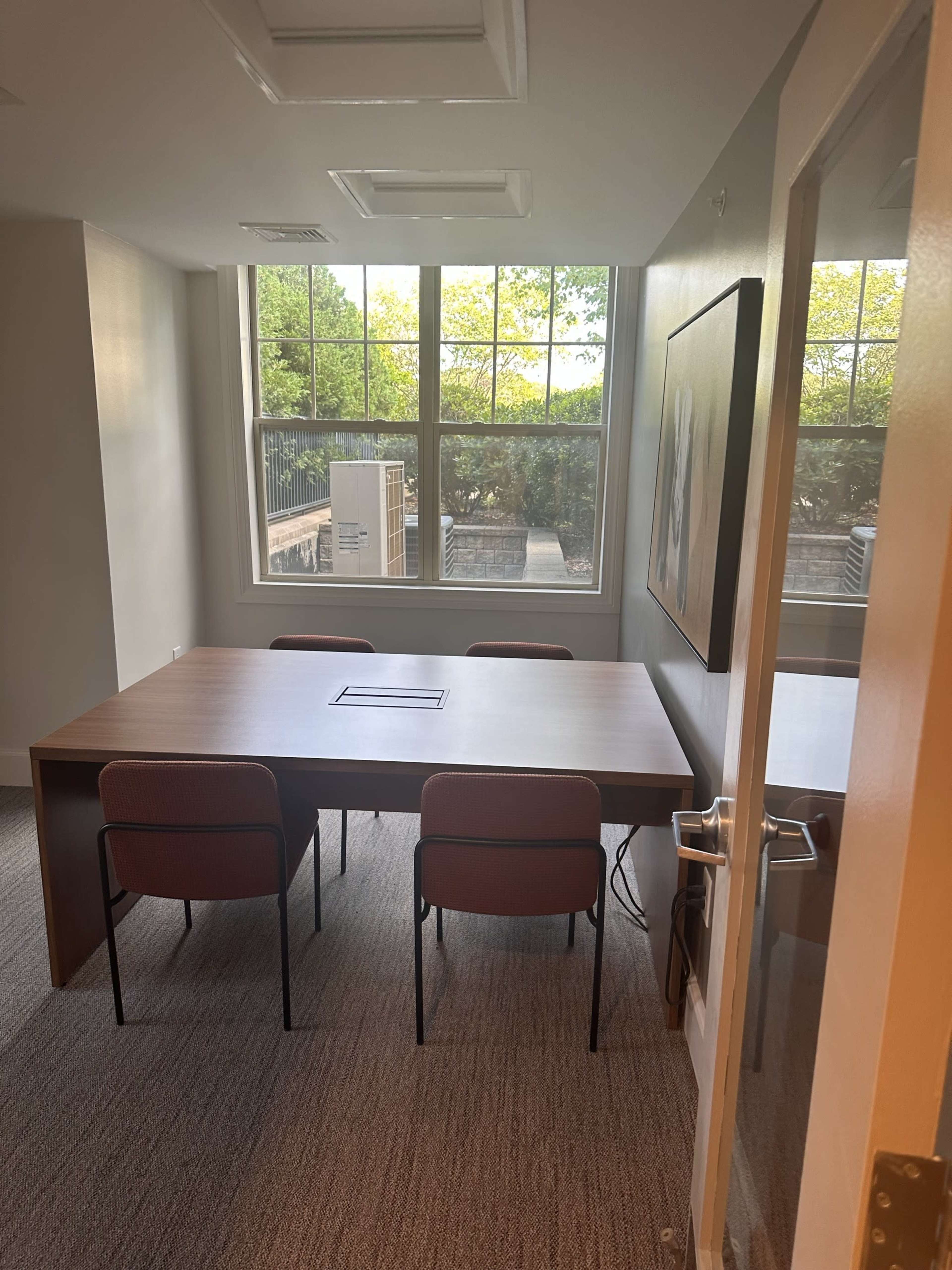 A conference room with a large table and four chairs, illuminated by natural light from a window.