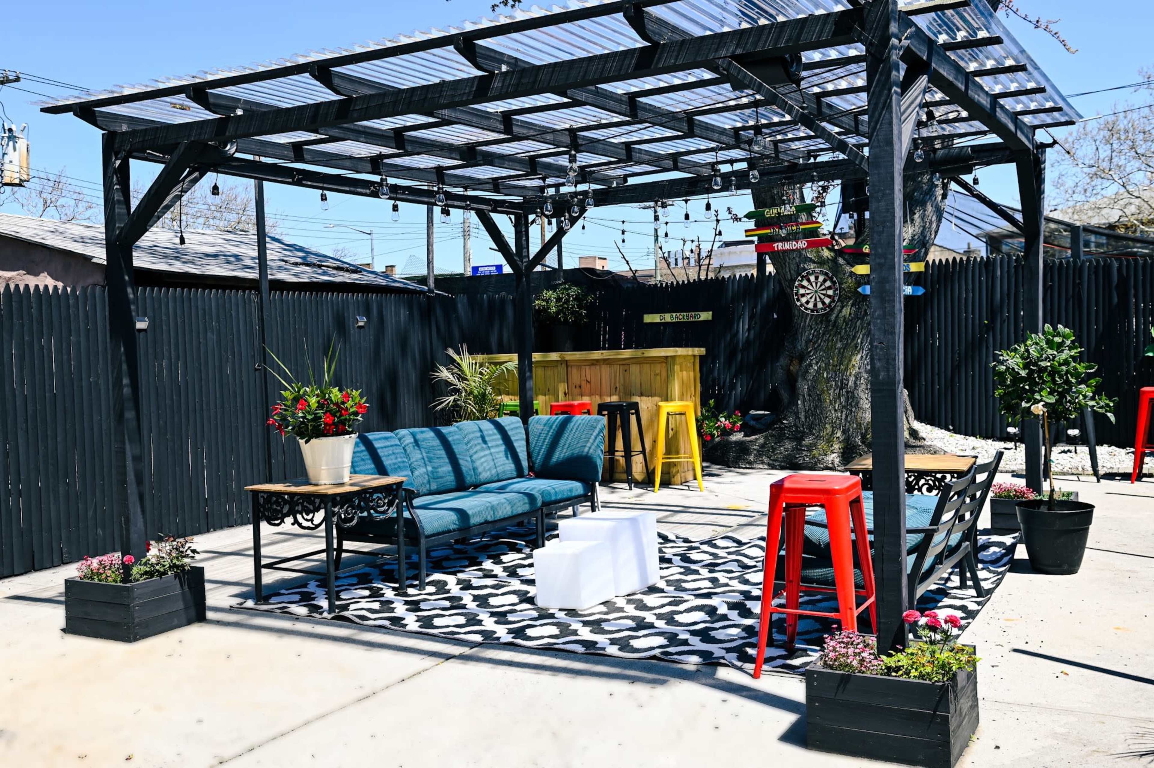A patio area with a covered seating arrangement, featuring blue sofas, a coffee table, and bright red stools, surrounded by decorative plants and a dartboard.