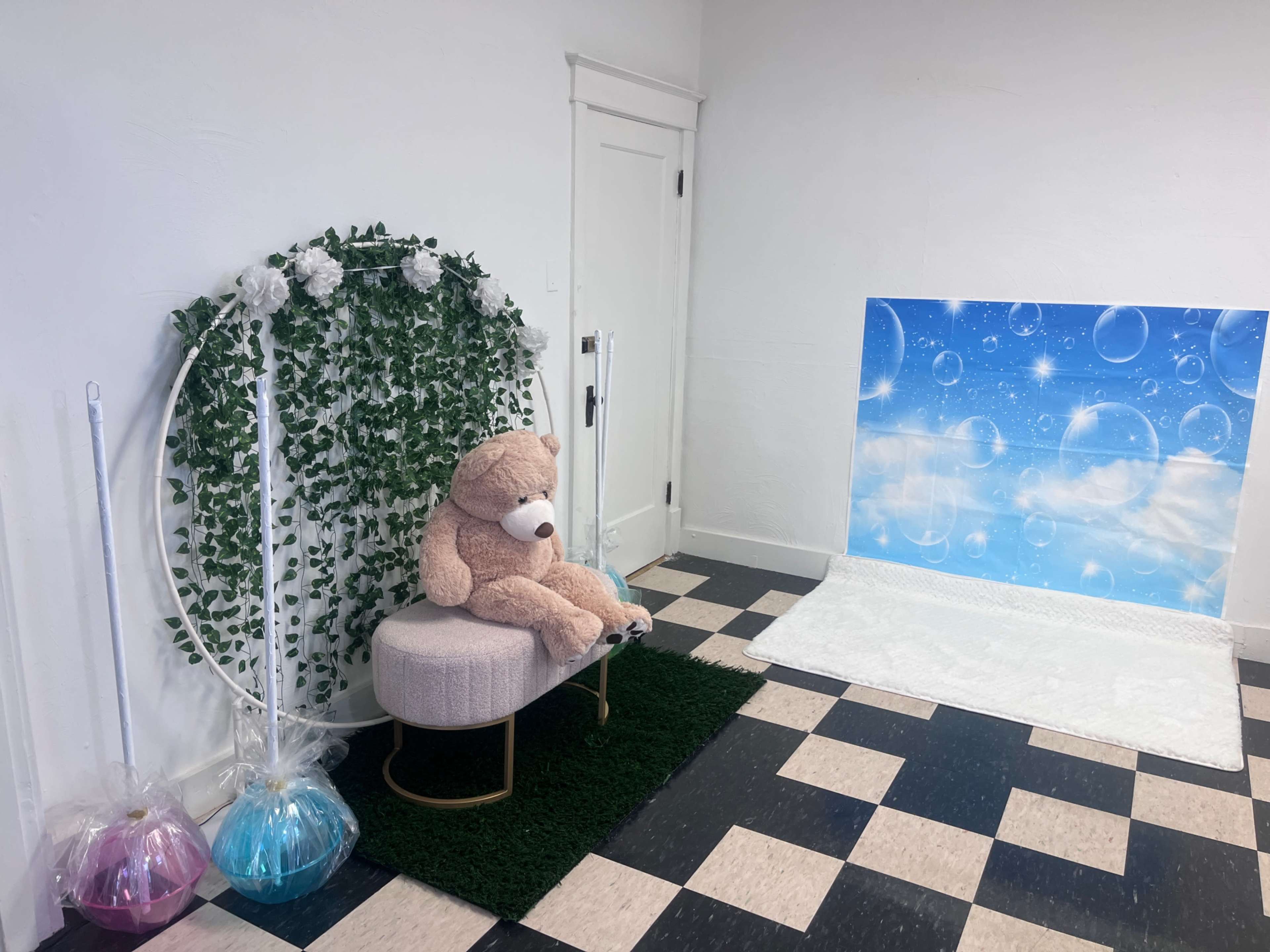 The image depicts a decorated indoor space featuring a large teddy bear sitting on a round, pink chair with greenery and white flowers in the background, alongside a backdrop with a blue sky and bubbles.