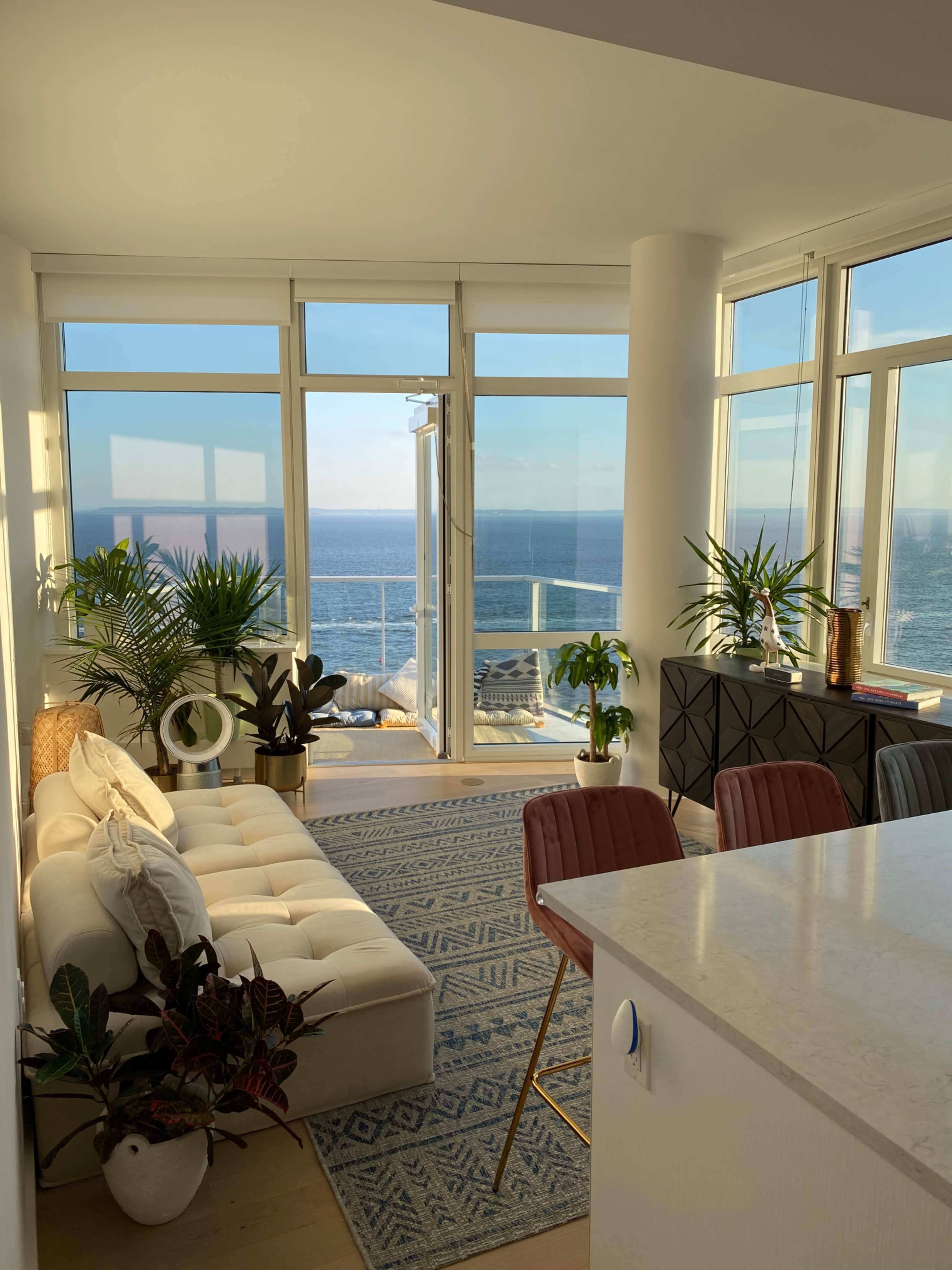 A bright living space features a plush sofa, indoor plants, and a large window overlooking the ocean.
