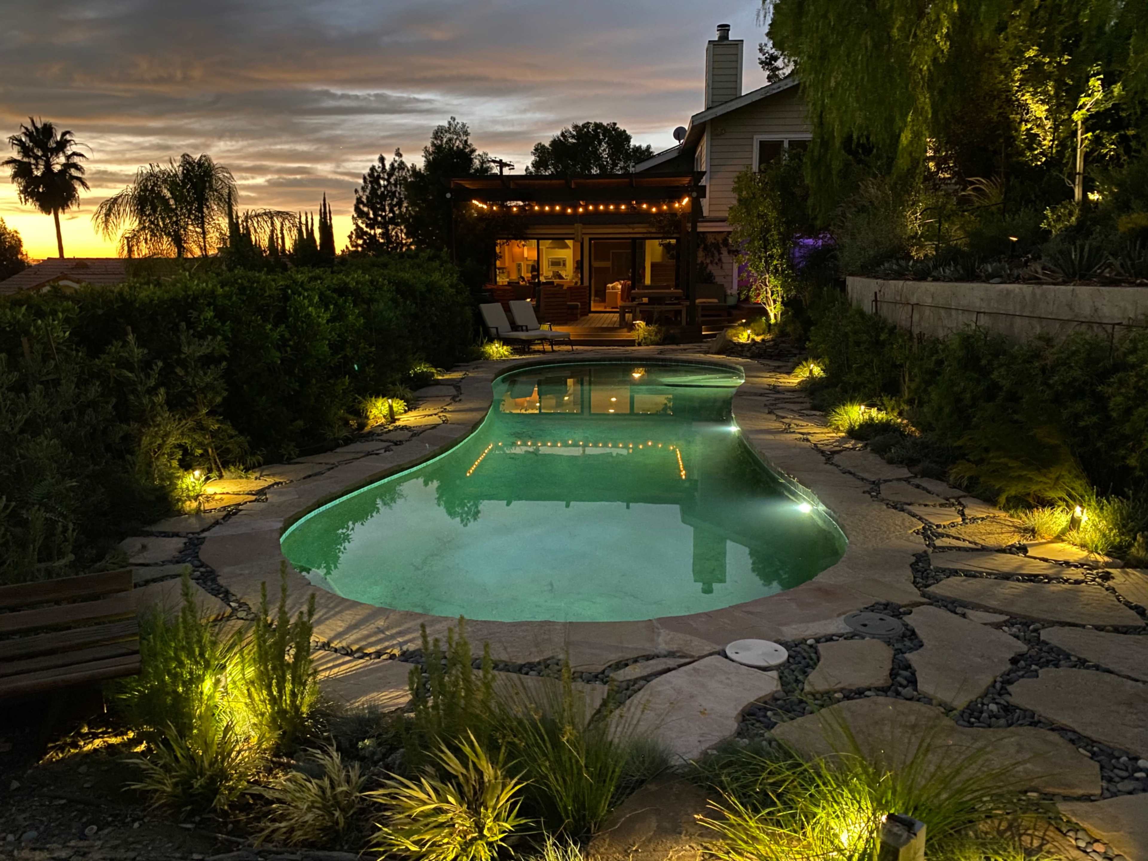 The image shows a well-lit swimming pool surrounded by lush greenery and stone pathways, with a house in the background under a sunset sky.