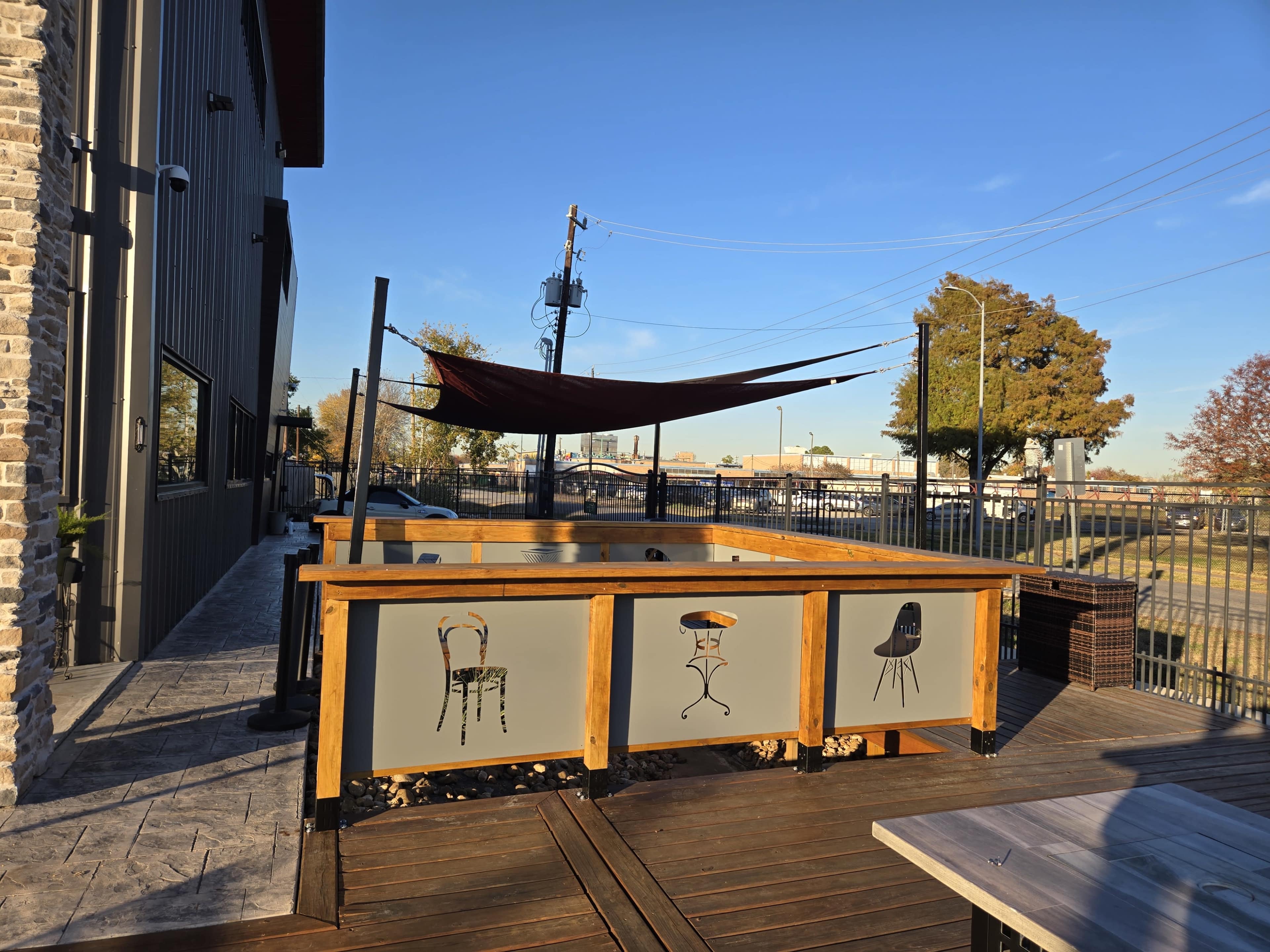 The image shows a wooden patio area with decorative chair illustrations on the railing and a shade sail above, set against a clear blue sky.