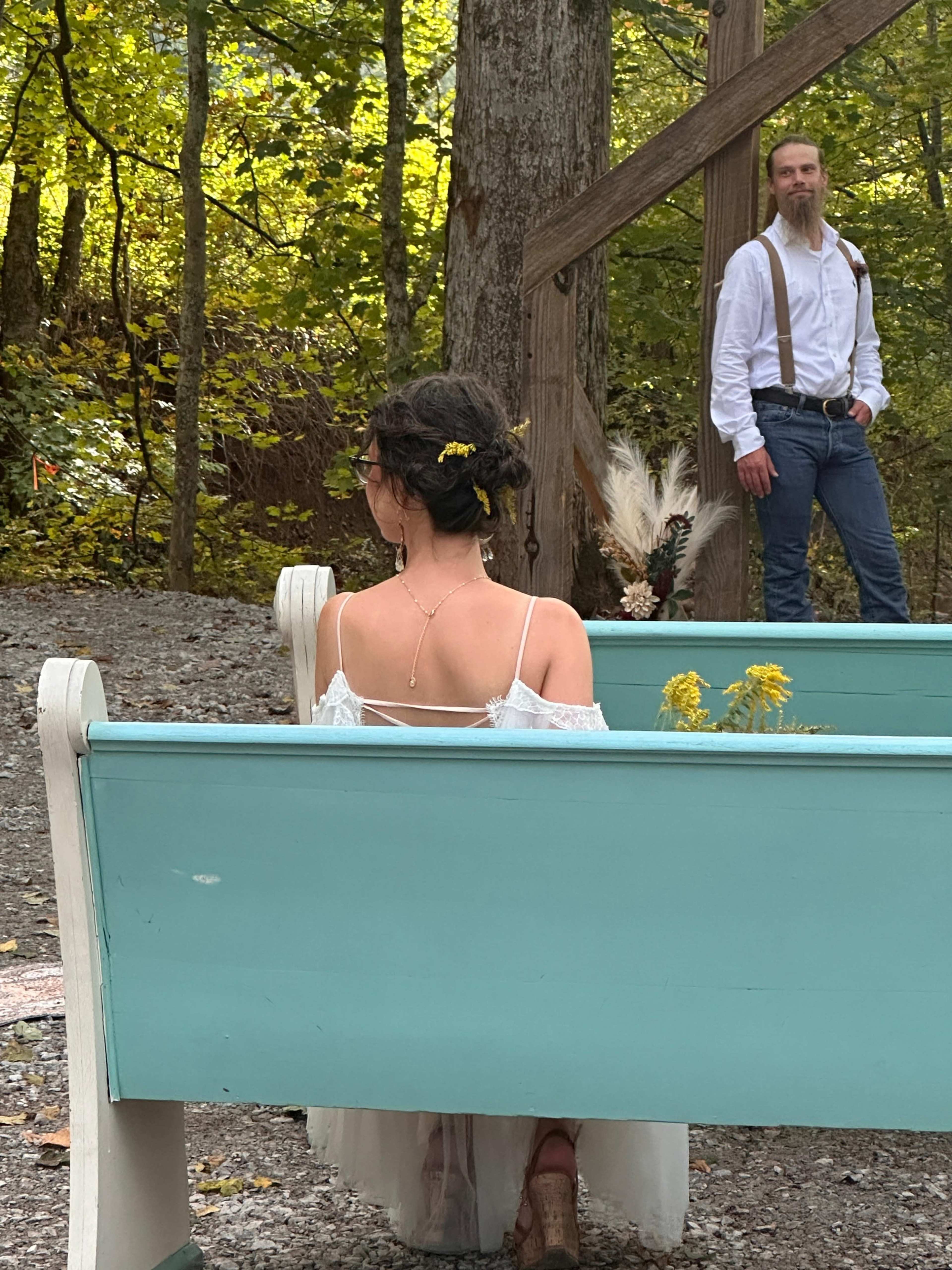 A woman in a white dress sits on a turquoise bench, while a man in a white shirt and suspenders stands in the background amid a wooded setting.