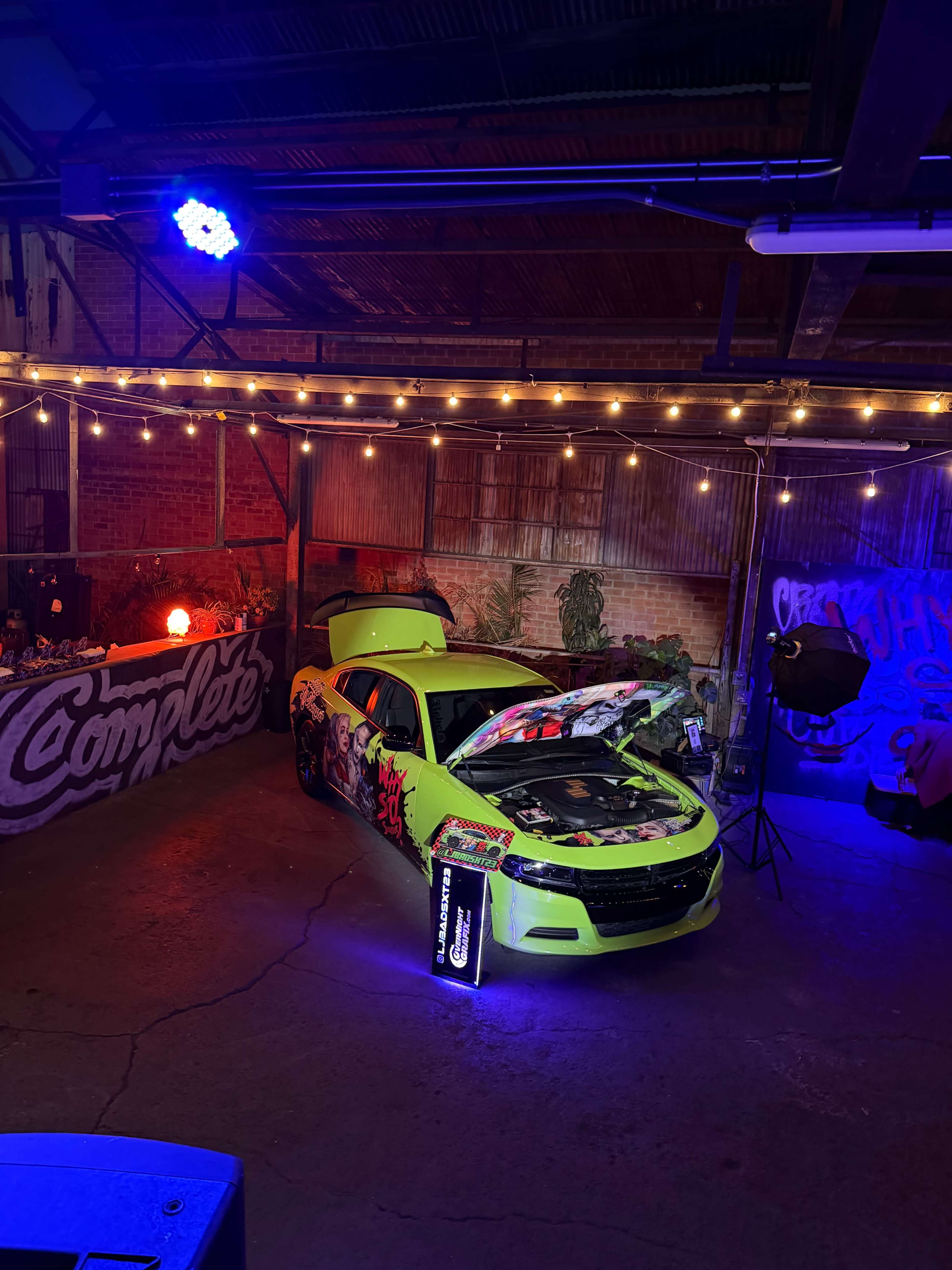 A brightly colored, custom-wrapped car with an open hood is displayed in an industrial-style venue adorned with string lights and graffiti.