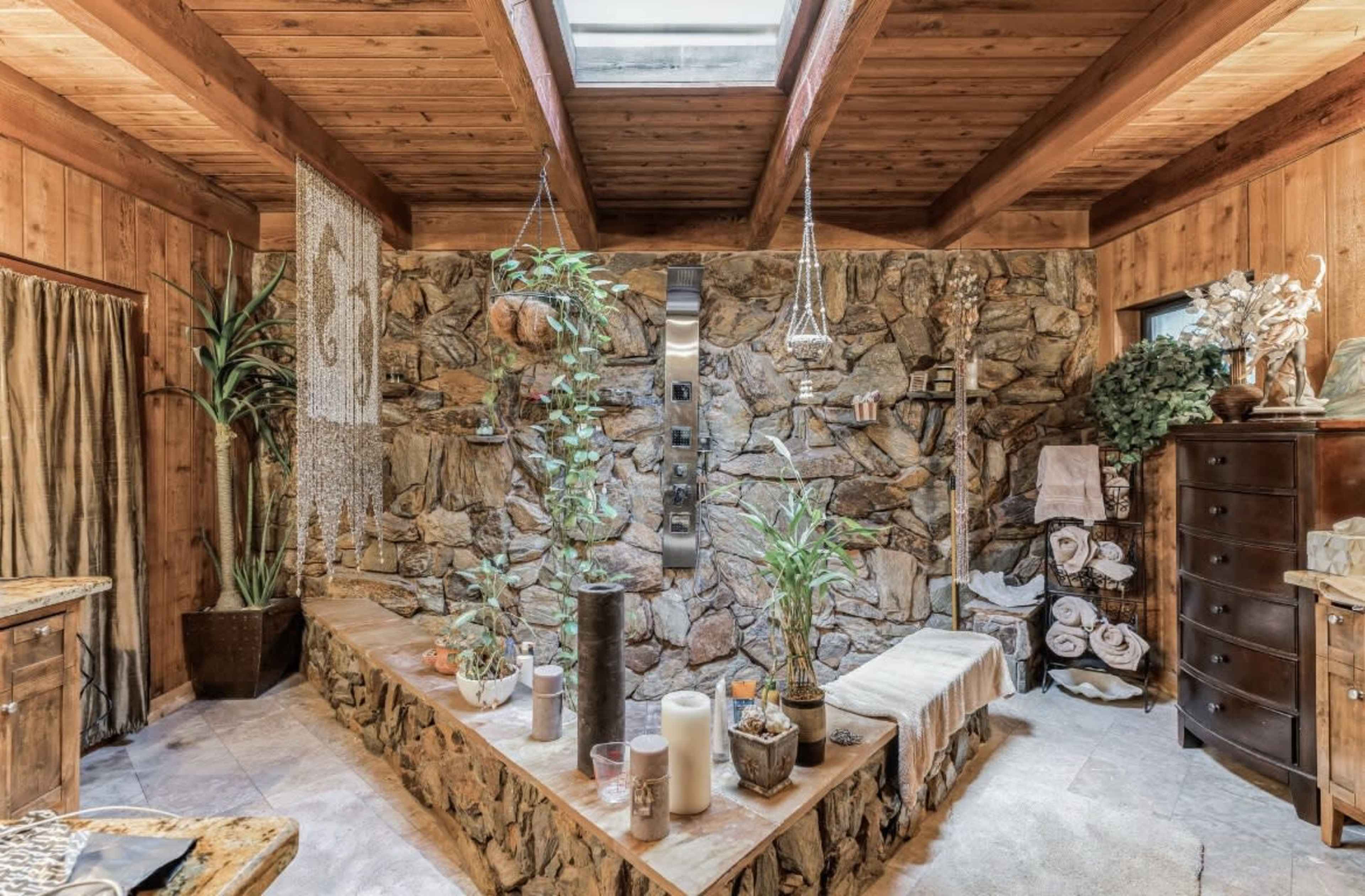 The image shows a rustic bathroom featuring a stone wall, wooden beams, and various plants arranged on a stone bench.