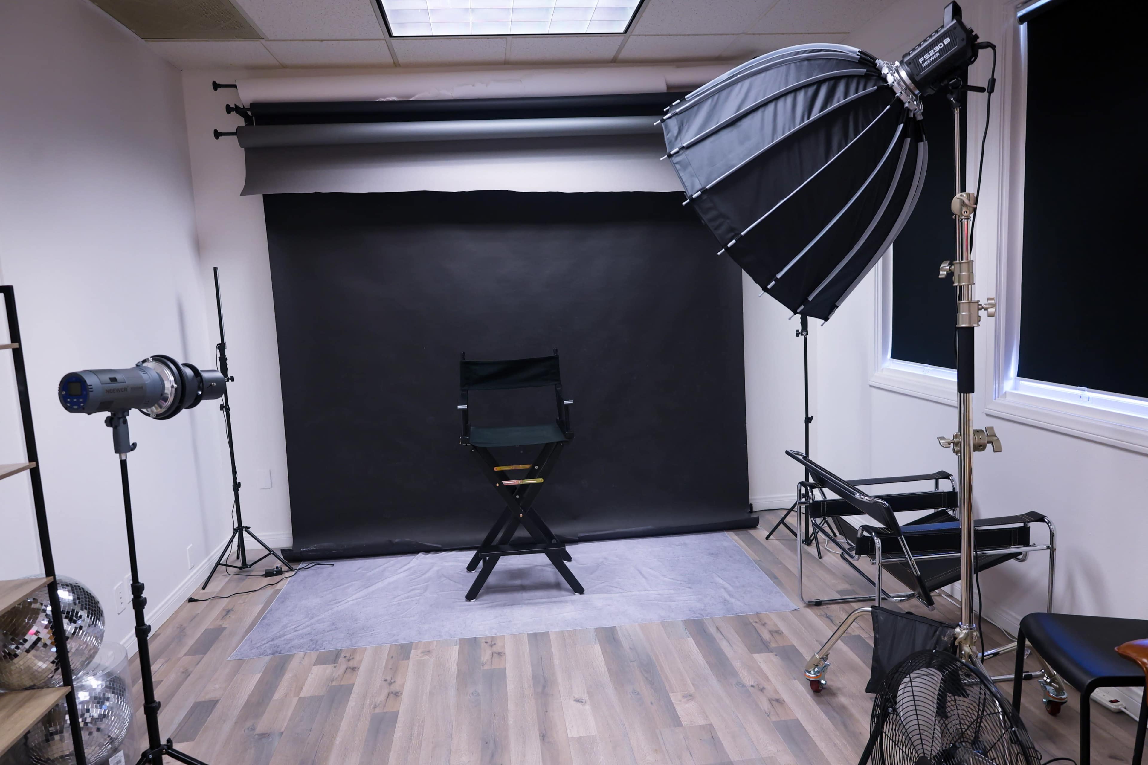 The image shows a photography studio equipped with a black backdrop, a director's chair, lighting equipment, and various props arranged neatly.