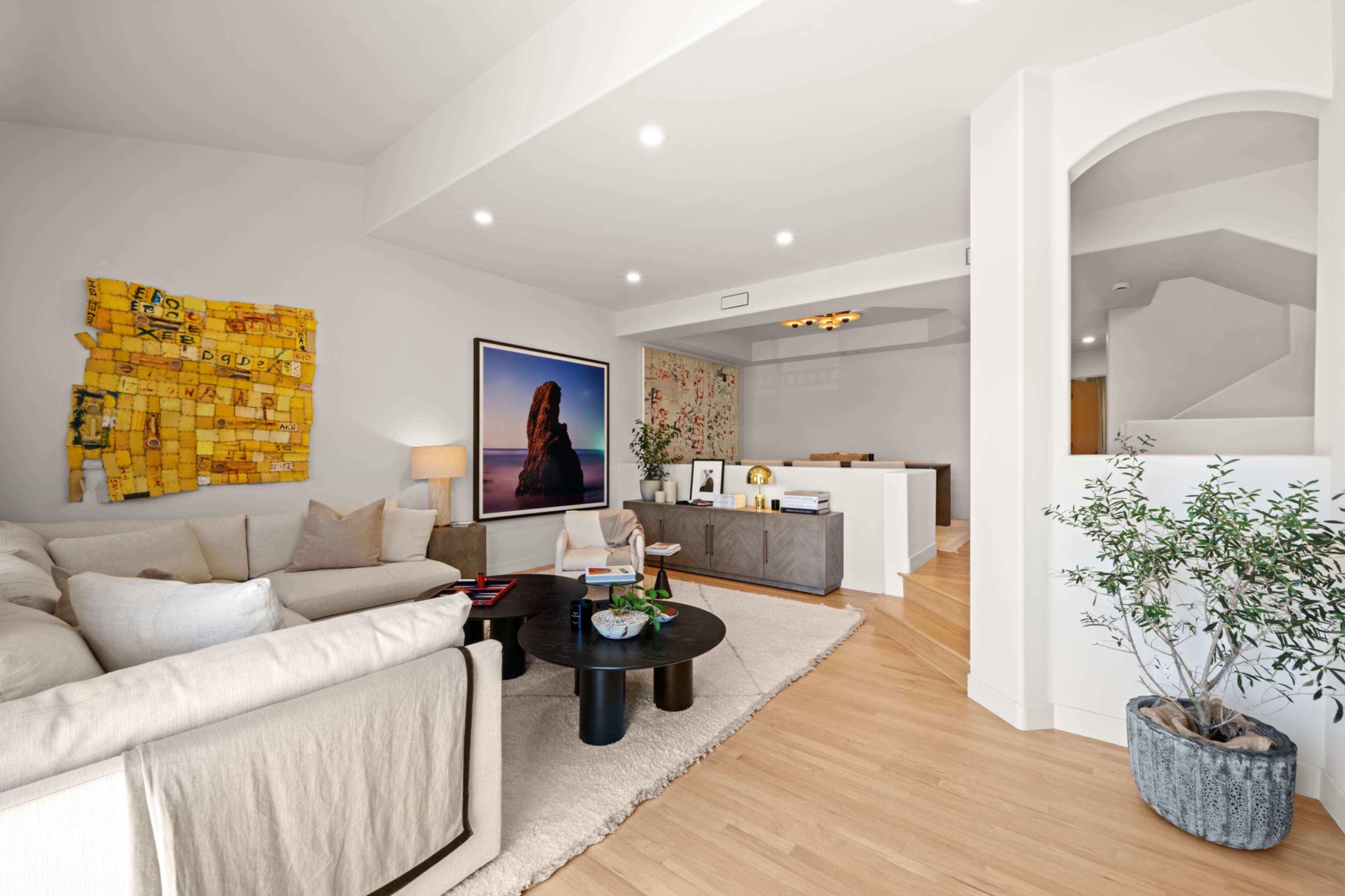 The image shows a modern living room with light wood flooring, a large beige sectional sofa, a circular black coffee table, and a wall decorated with art and a large framed photo.