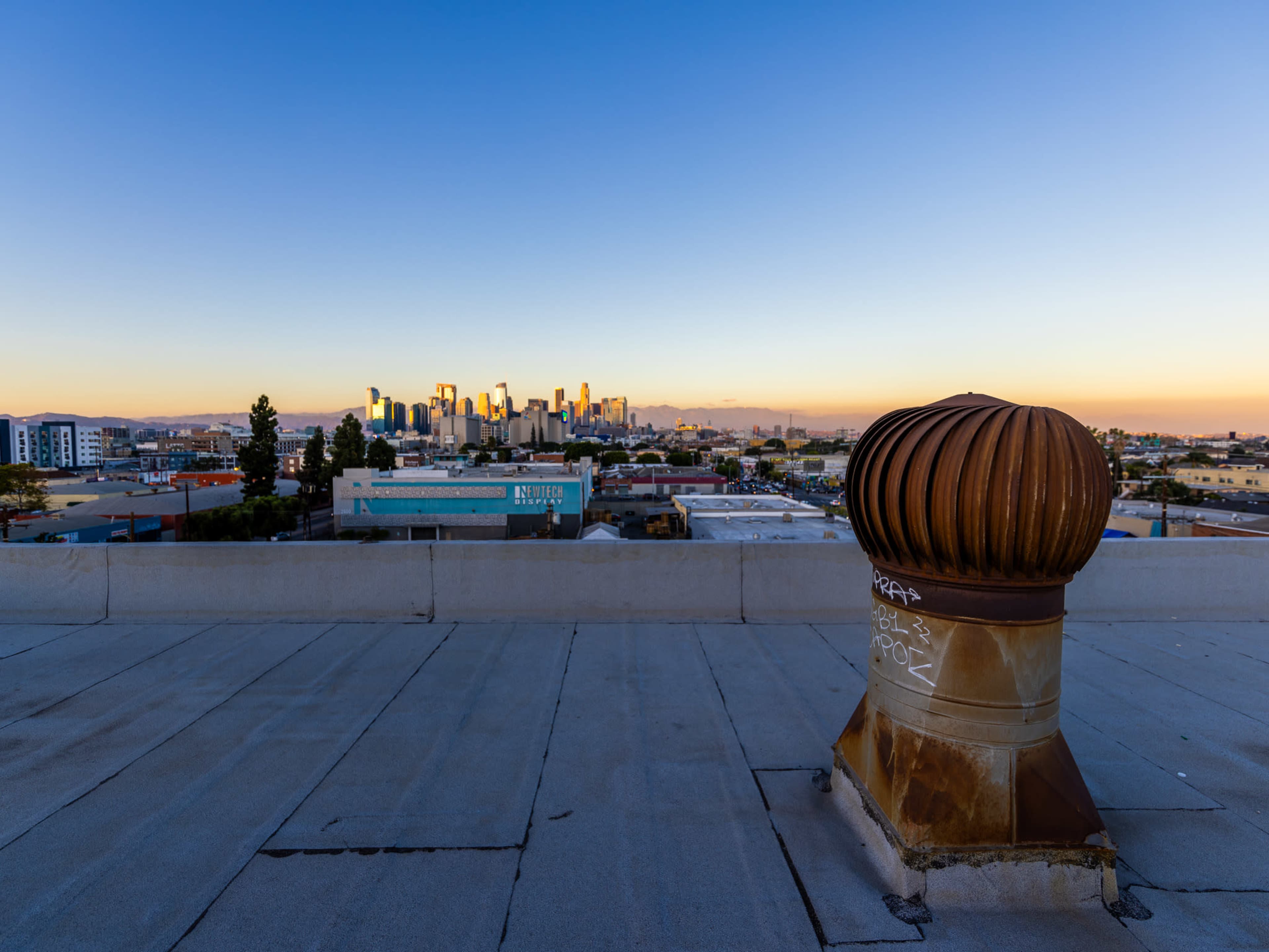 A rooftop ventilation fan is positioned in the foreground with the skyline of a city visible in the background during sunset.