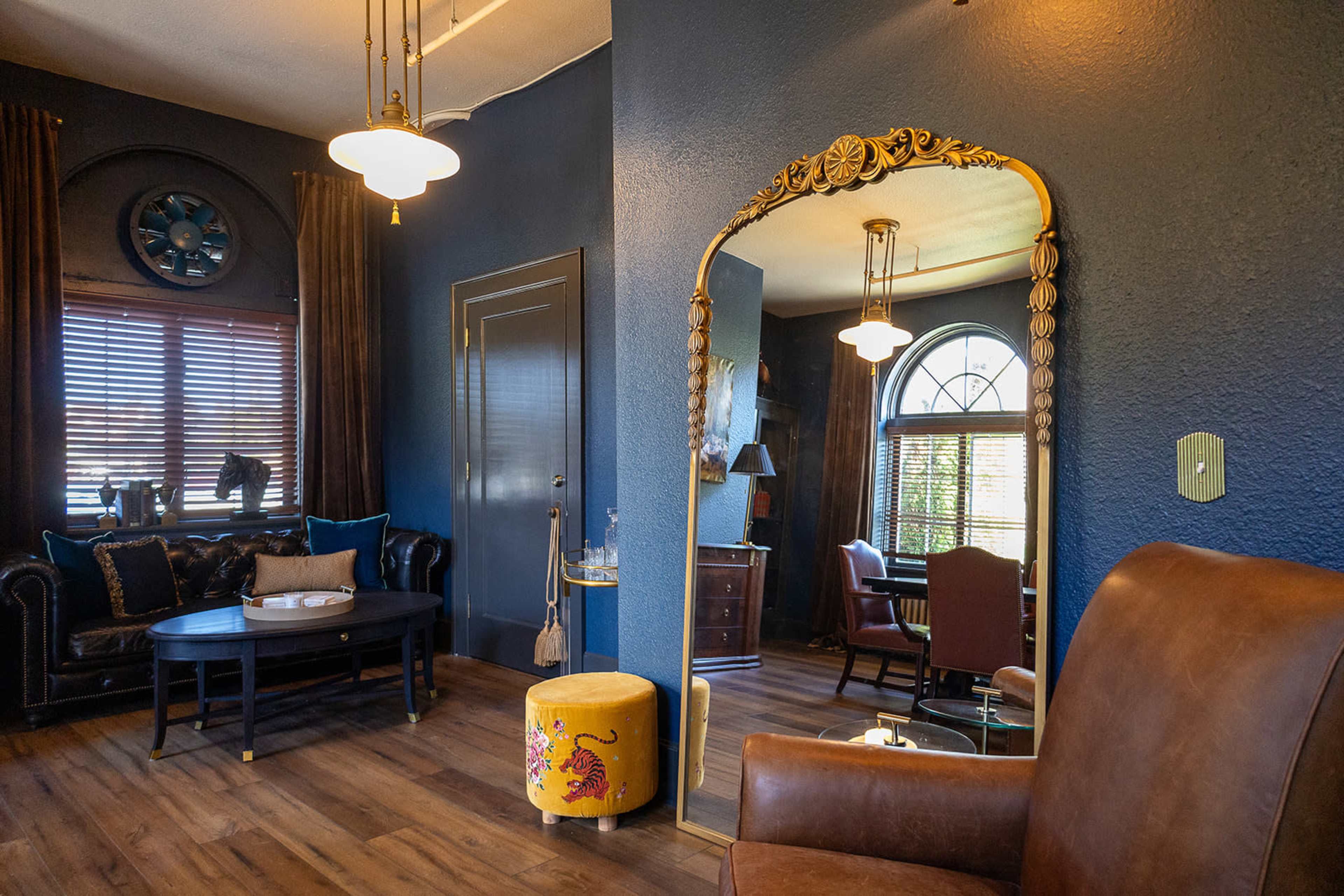 The image shows a stylish room with dark blue walls, a large ornate mirror, a black couch, a circular clock, and a mix of wooden and upholstered furniture.