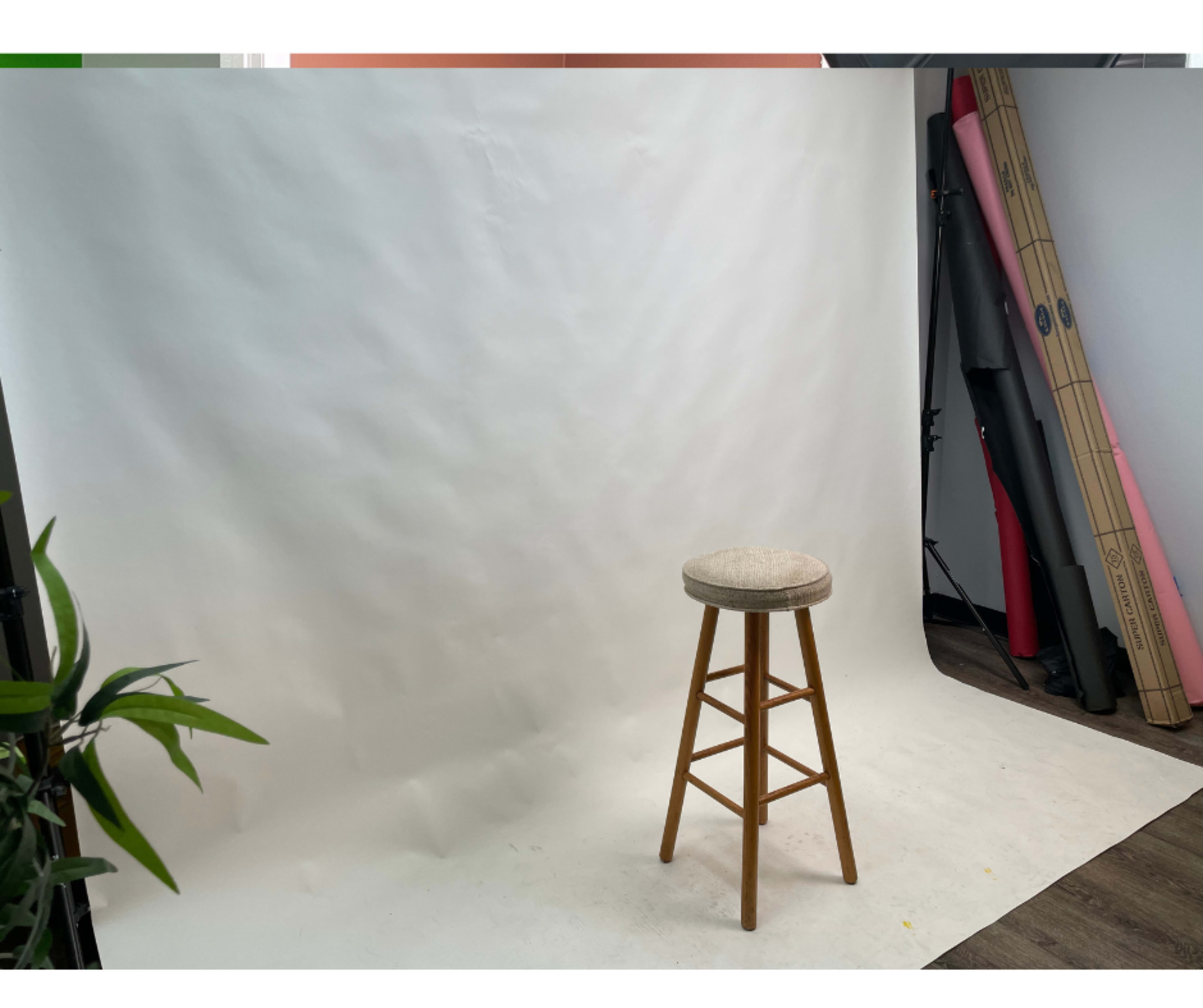 A plain white backdrop with a wooden stool positioned off-center and some photography equipment visible in the background.